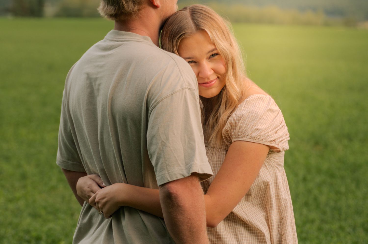 Young couple embracing in a sunny green field, woman smiling over her shoulder.