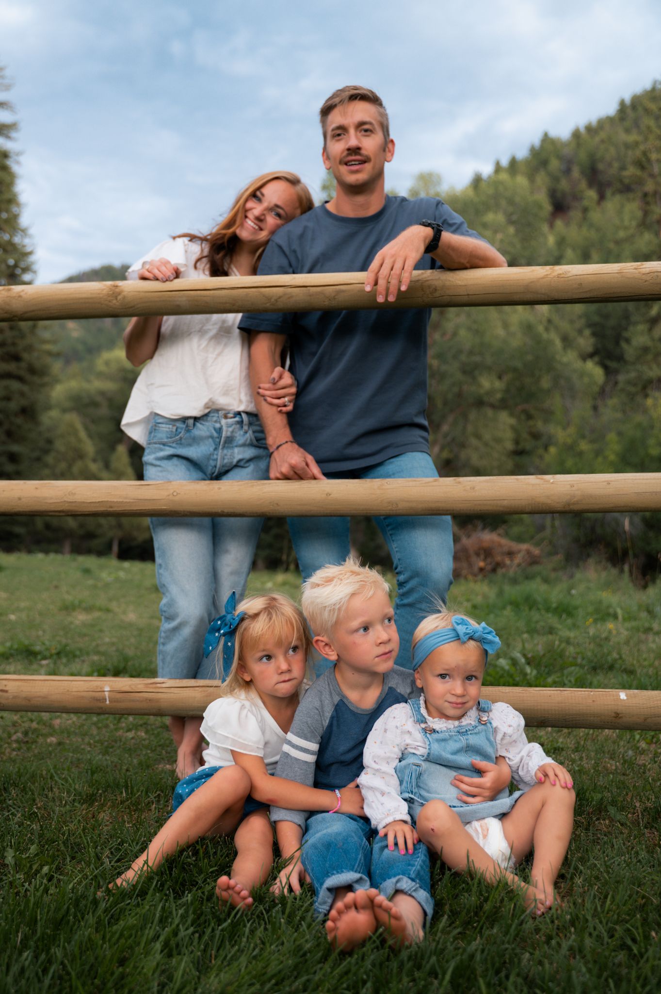Family portrait with couple leaning on fence and three young children sitting on grass.