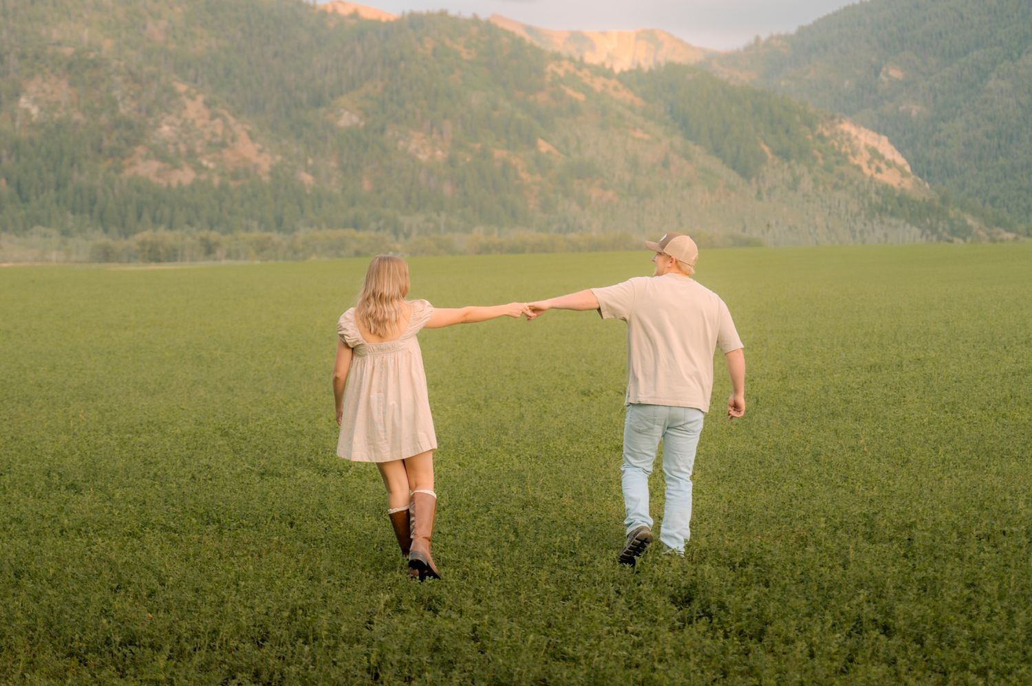 Couple holding hands walking through a green field with mountain backdrop at golden hour.
