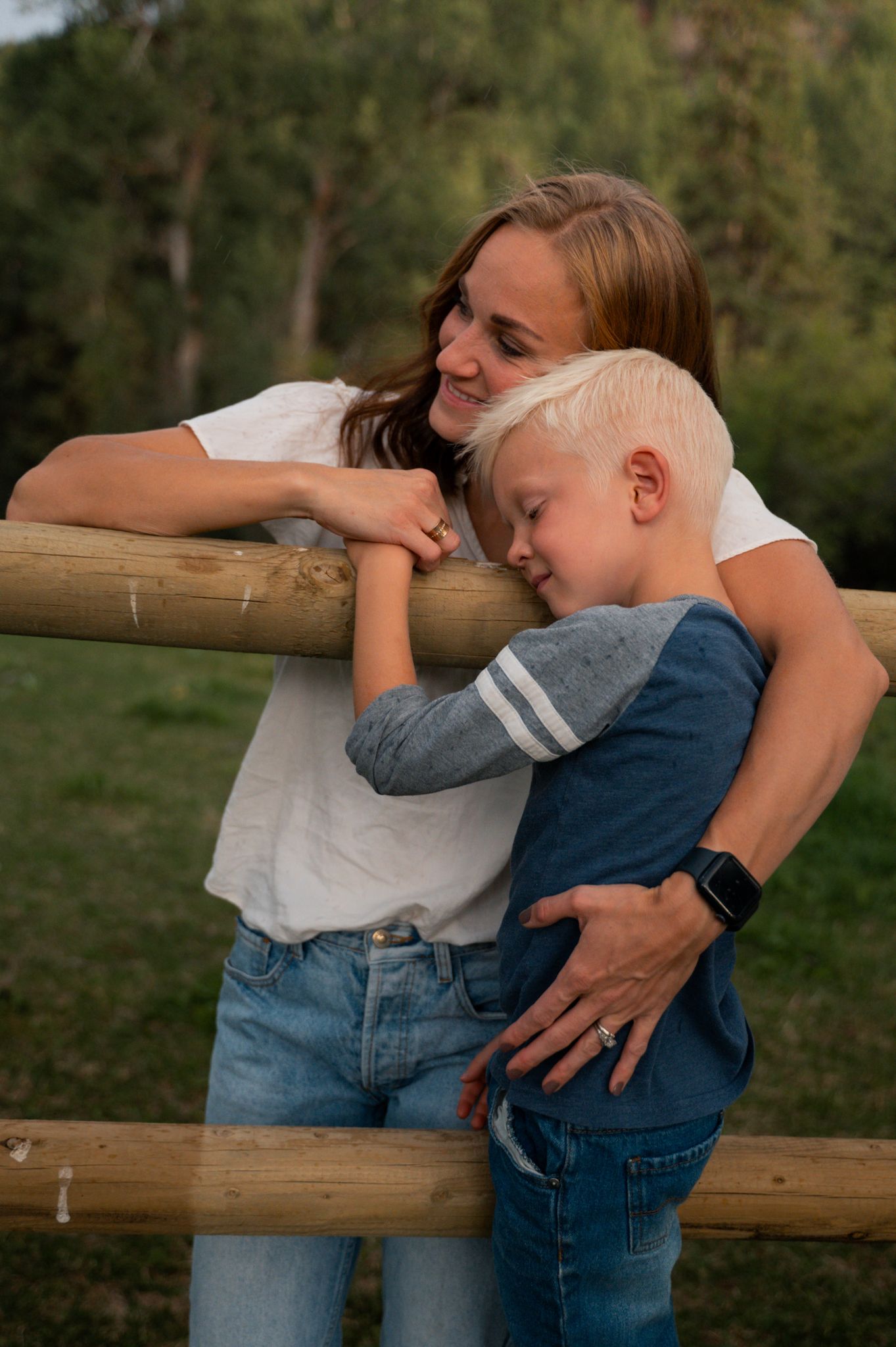 Mother and young blonde boy share a tender moment leaning against a rustic wooden fence outdoors.