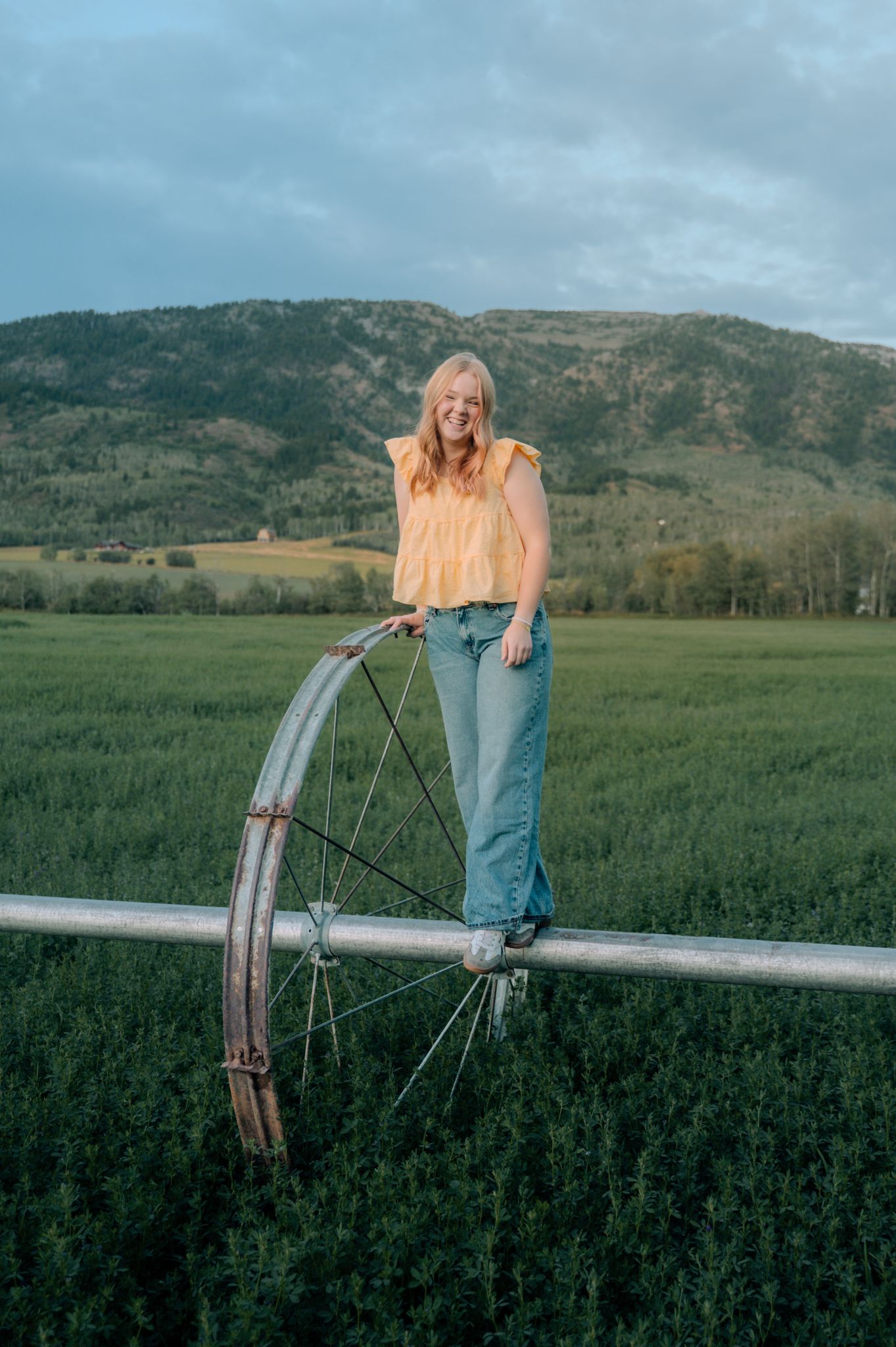 Young woman in yellow top and jeans standing on farm irrigation equipment in a green field with mountain backdrop.