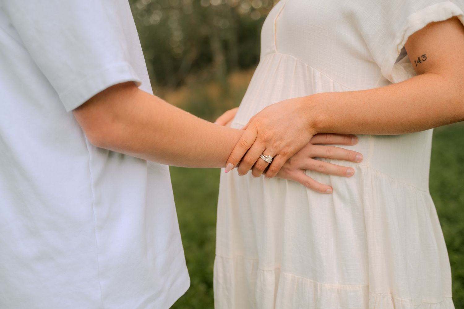 Close-up of couple holding hands on pregnant belly, woman wearing white dress and diamond ring outdoors.