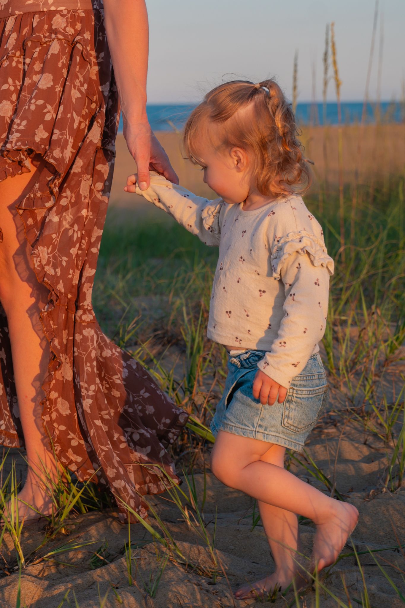 Toddler girl in denim skirt holding mothers hand walking barefoot on sandy beach.