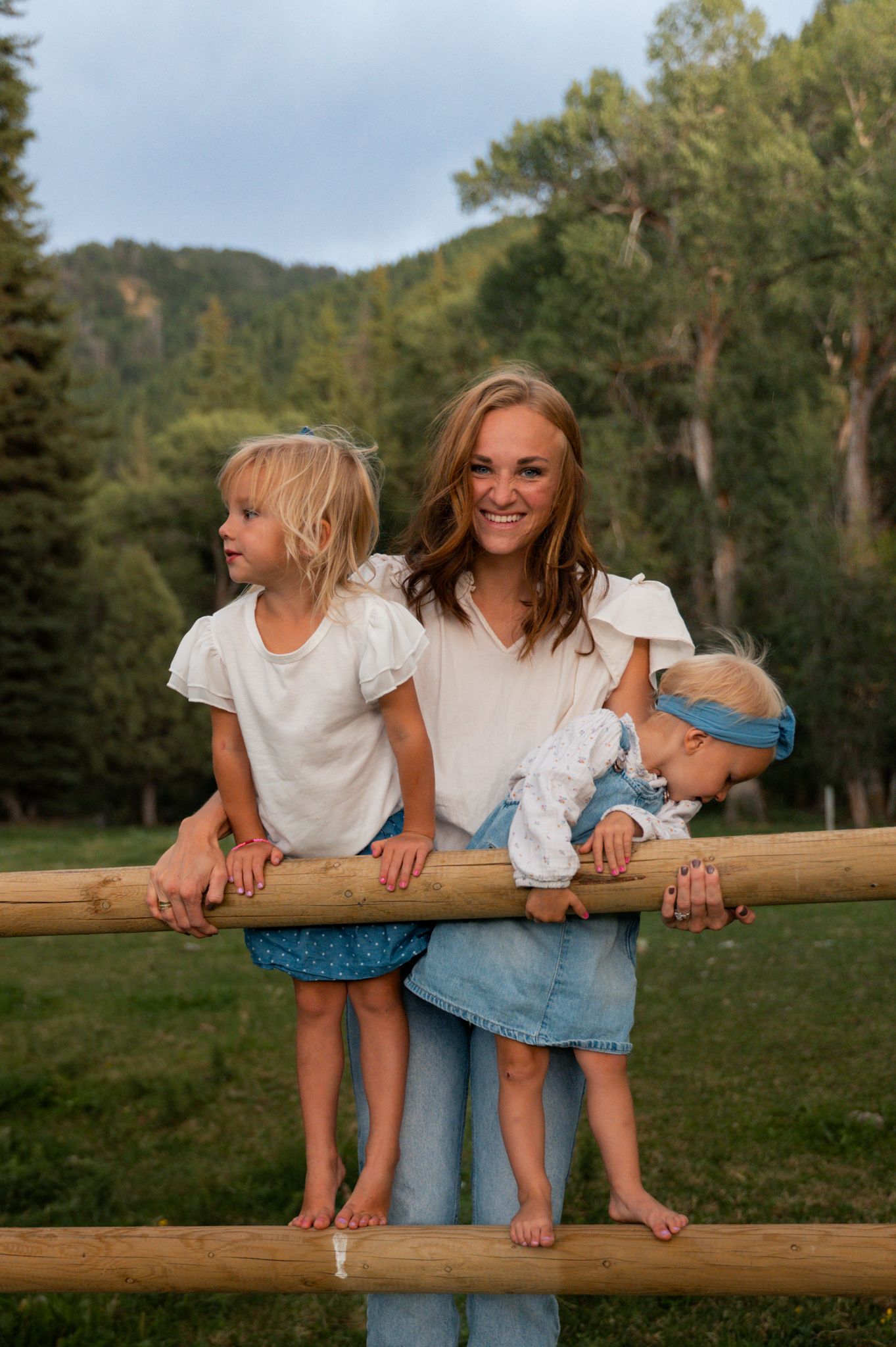 Smiling mother with two young daughters leaning on a rustic wooden fence outdoors.