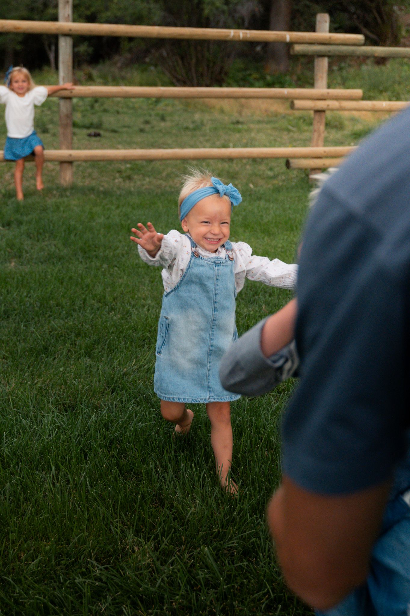 Happy little girl in denim dress and blue bow running across green grass outdoors.