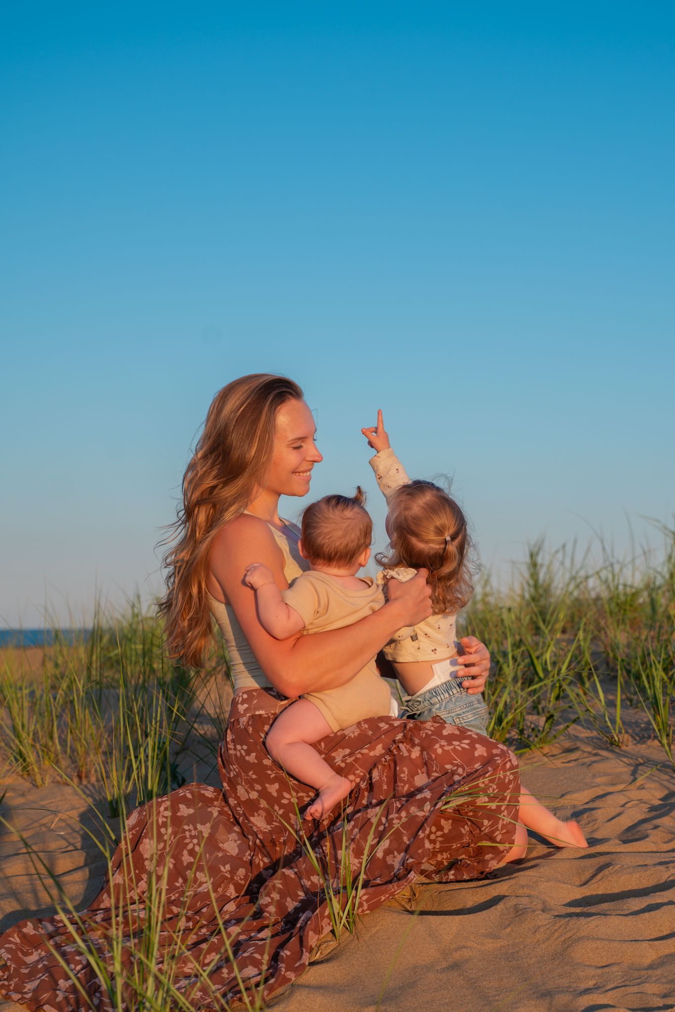 A smiling mother sits on a beach dune holding a newborn while her toddler points toward the sky.