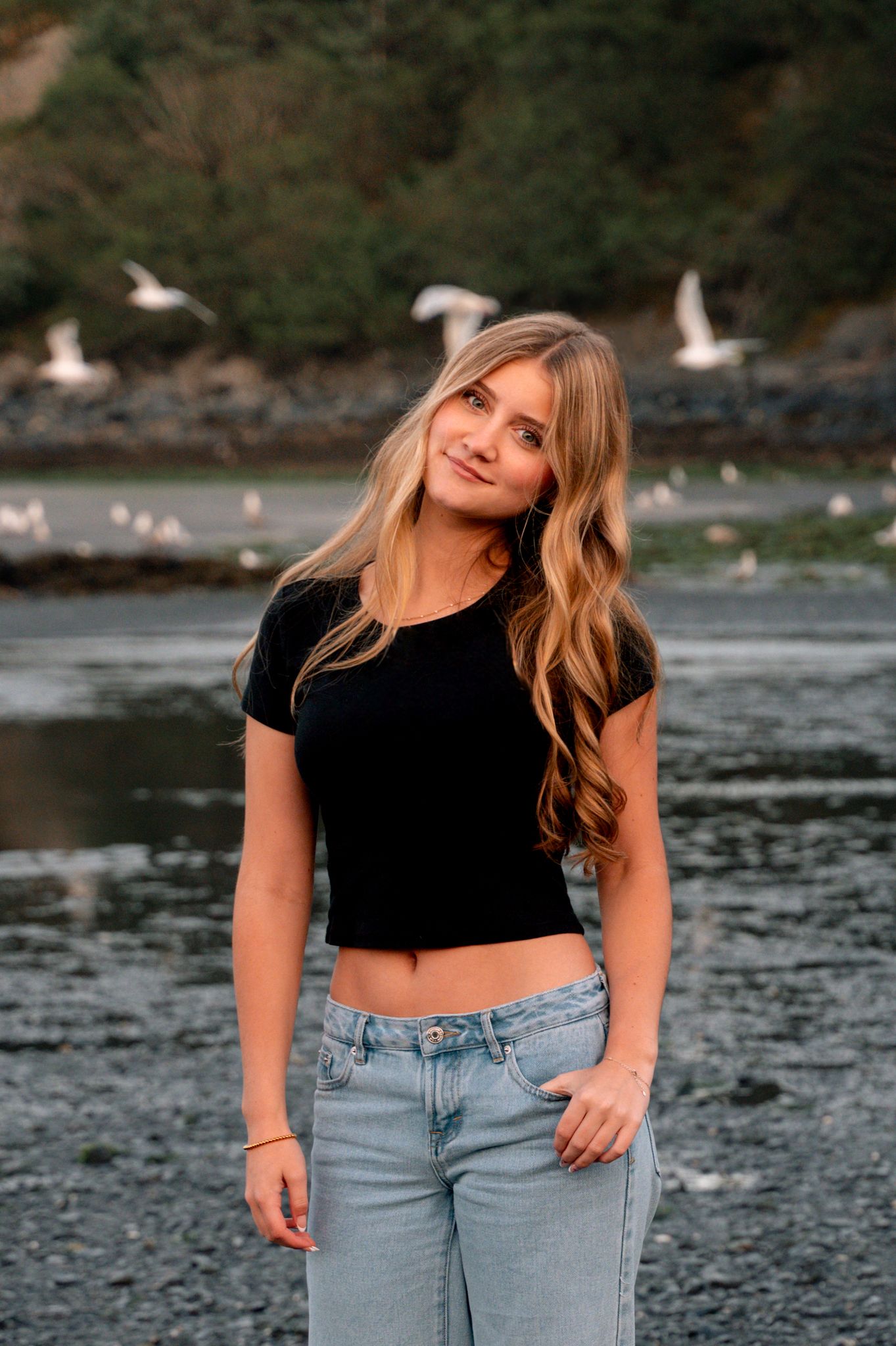 Young blonde woman in black crop top and light jeans poses on rocky beach with seagulls flying in background.