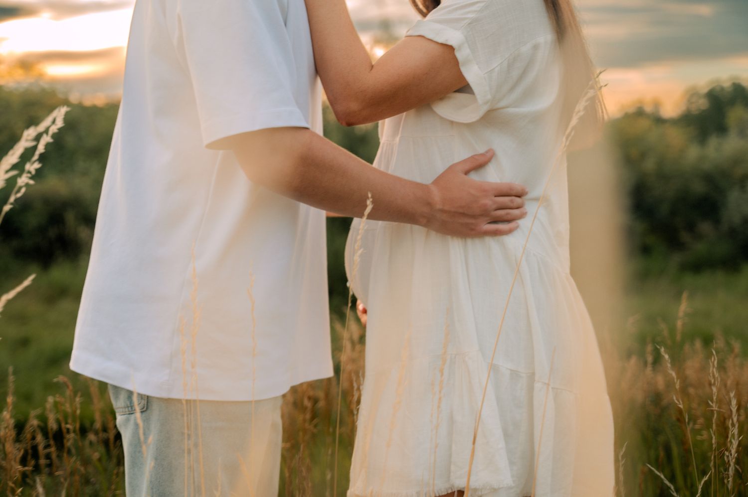 Couple in white outfits embracing in a sunlit field of tall grass at golden hour.