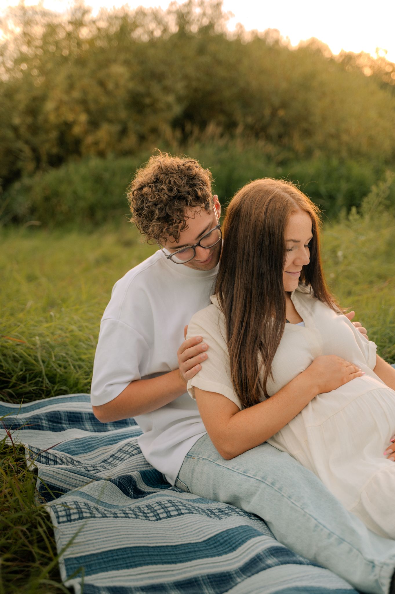 Young couple sitting on blanket in green field at sunset, man embracing pregnant woman from behind.
