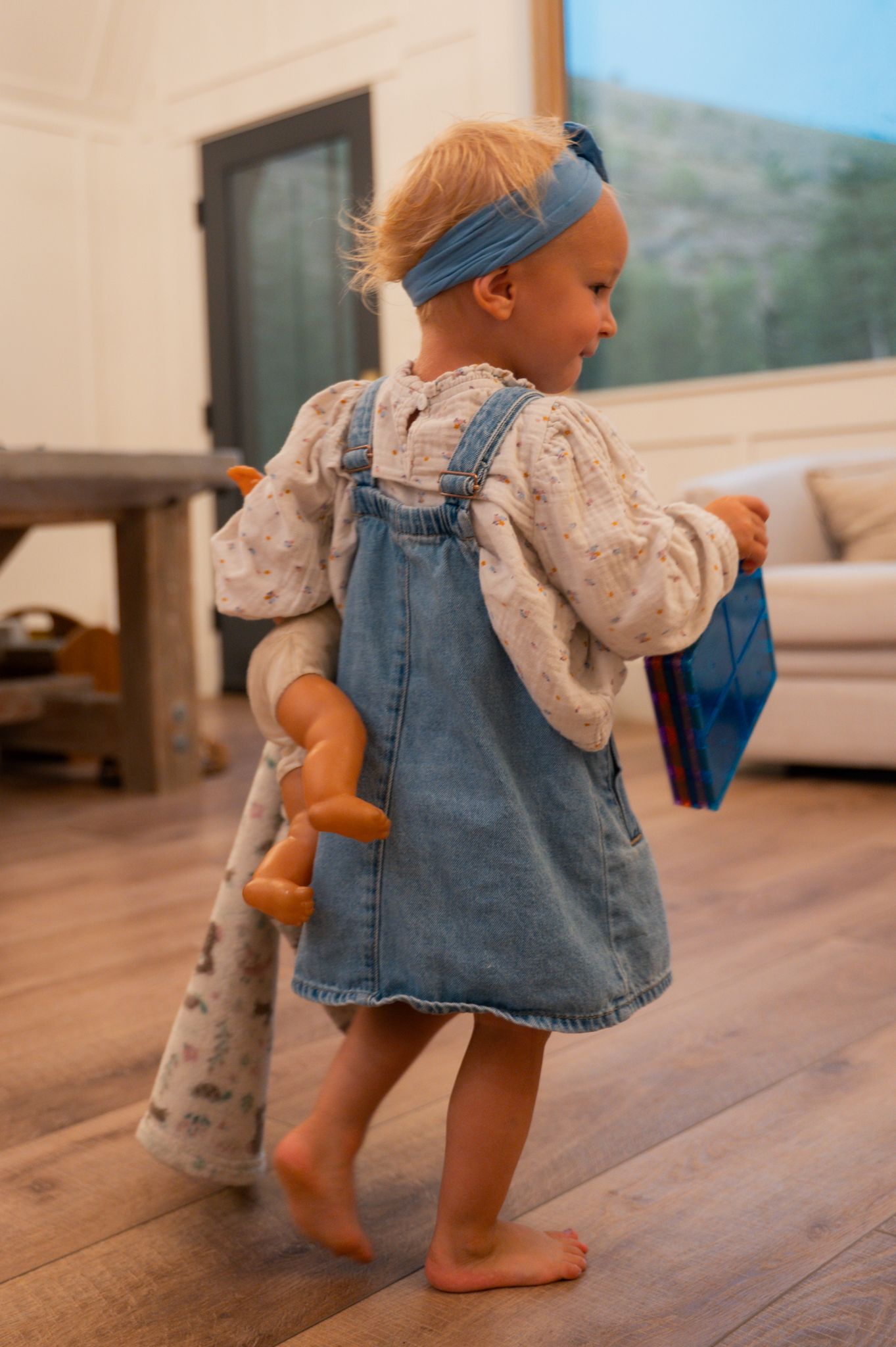 Toddler girl in denim overalls walks indoors carrying a doll and small blue book on wood floors.