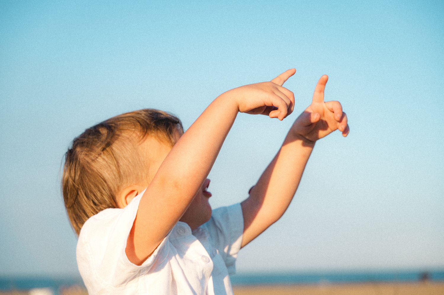 Young toddler in white shirt pointing fingers up toward the sky on a sunny beach.