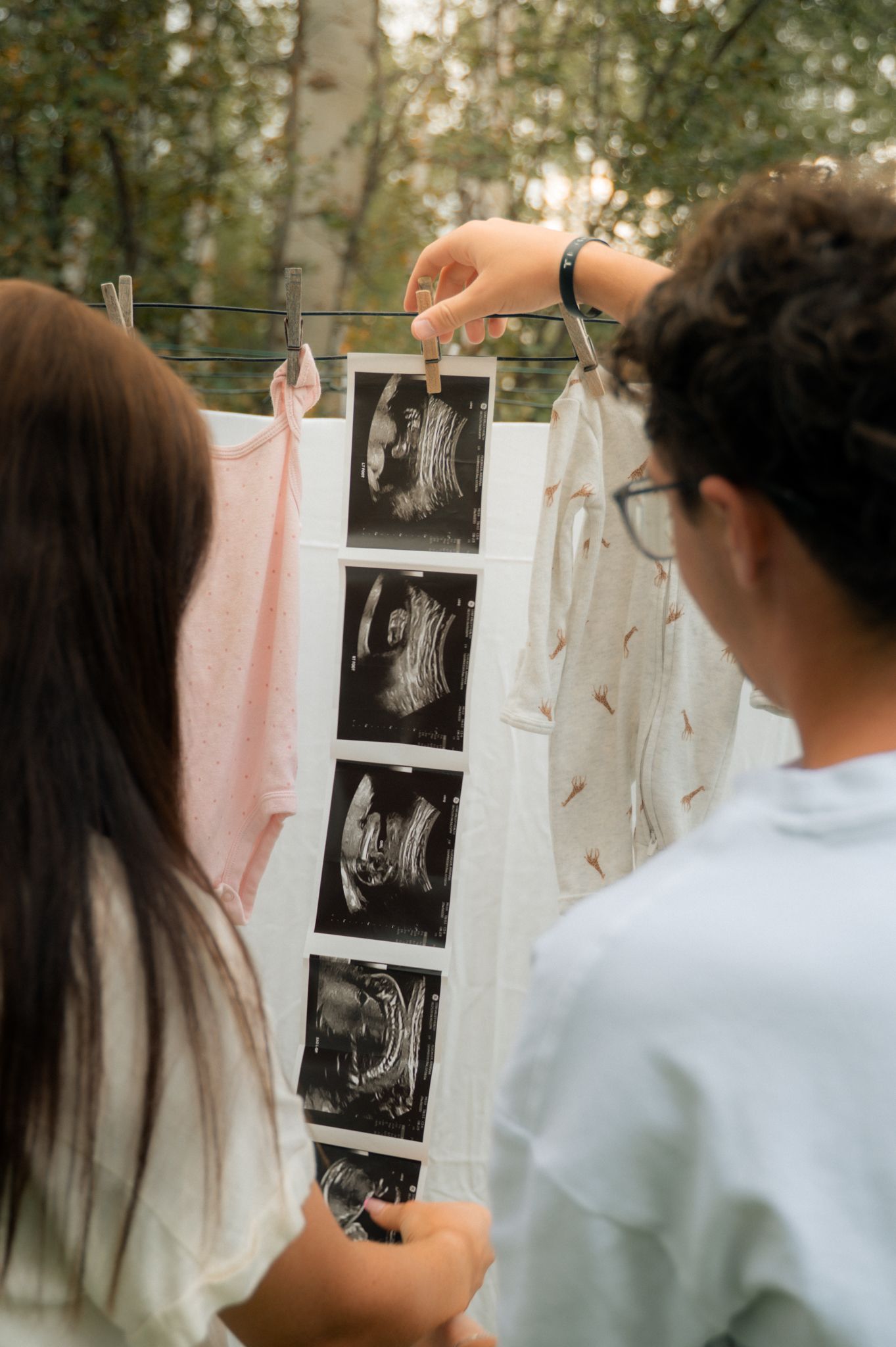 Couple viewing ultrasound photos hanging on clothesline with baby clothes outdoors.