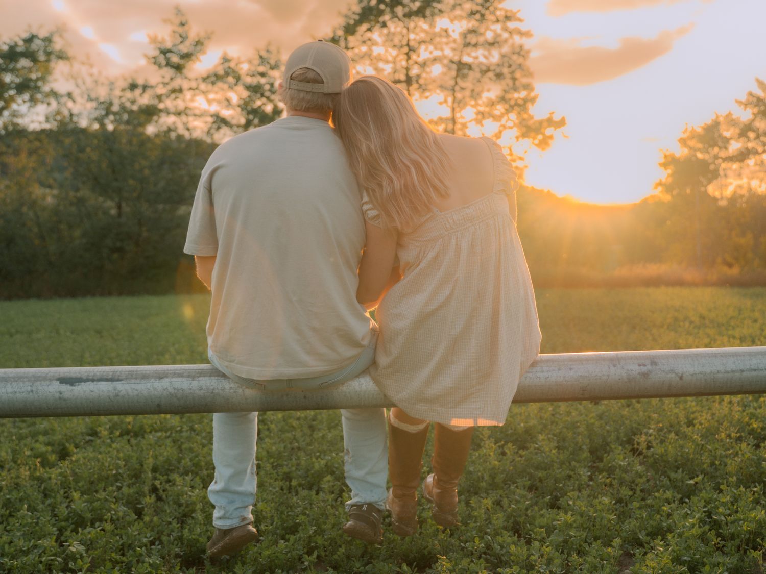 A couple sits together on a white fence rail watching a beautiful golden sunset over a lush green field.