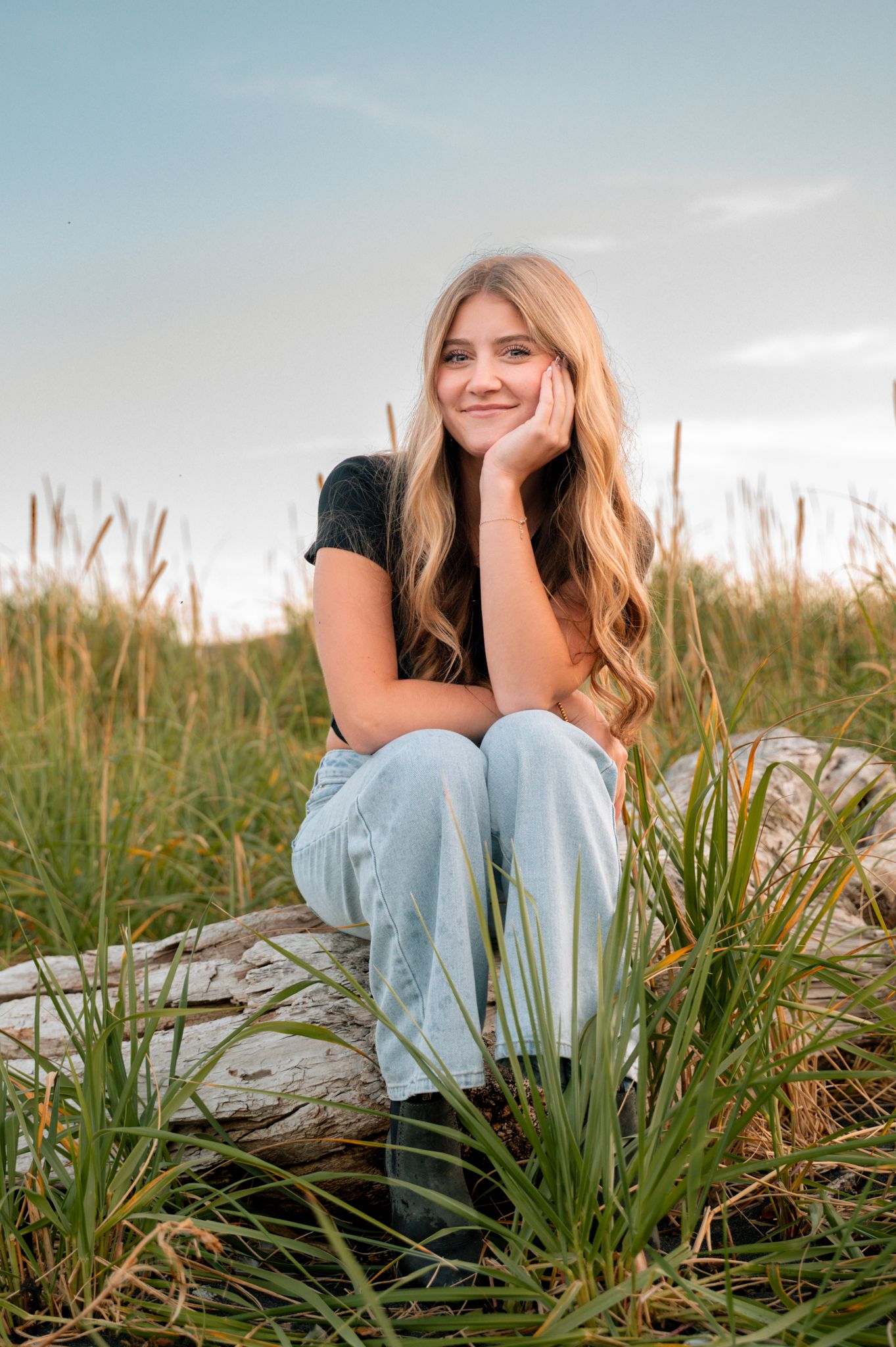 Young woman with long blonde hair sitting on driftwood in a grassy field, smiling warmly in jeans and black top.