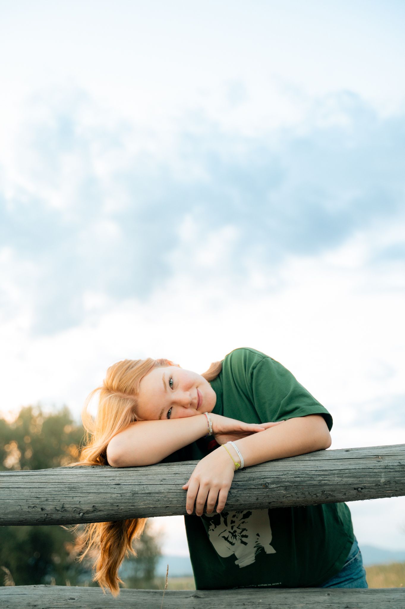 A red-haired teenage girl rests her head on a wooden fence rail under a cloudy sky.
