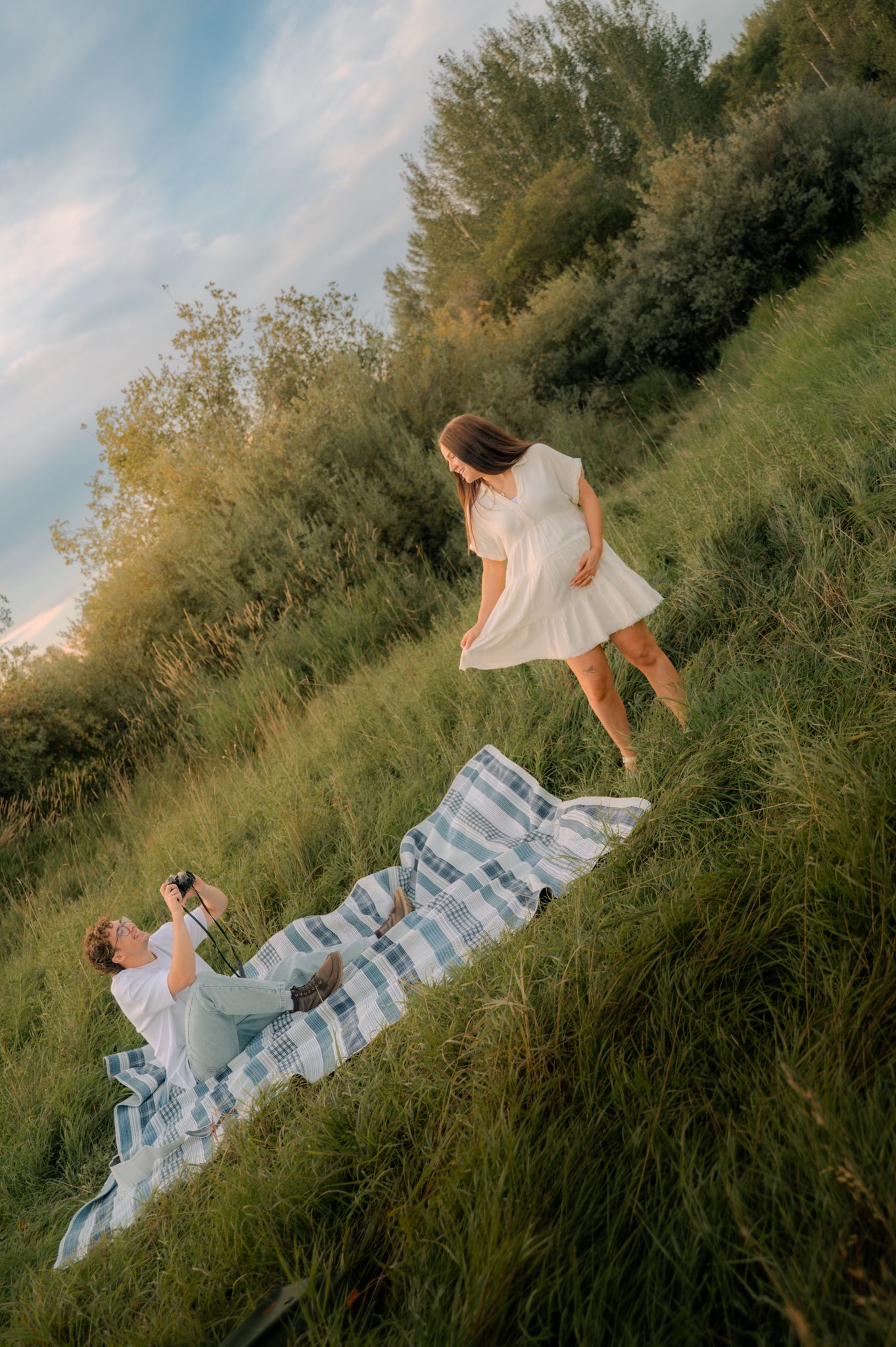 Pregnant woman in white dress standing on grassy hill while partner photographs her from below.