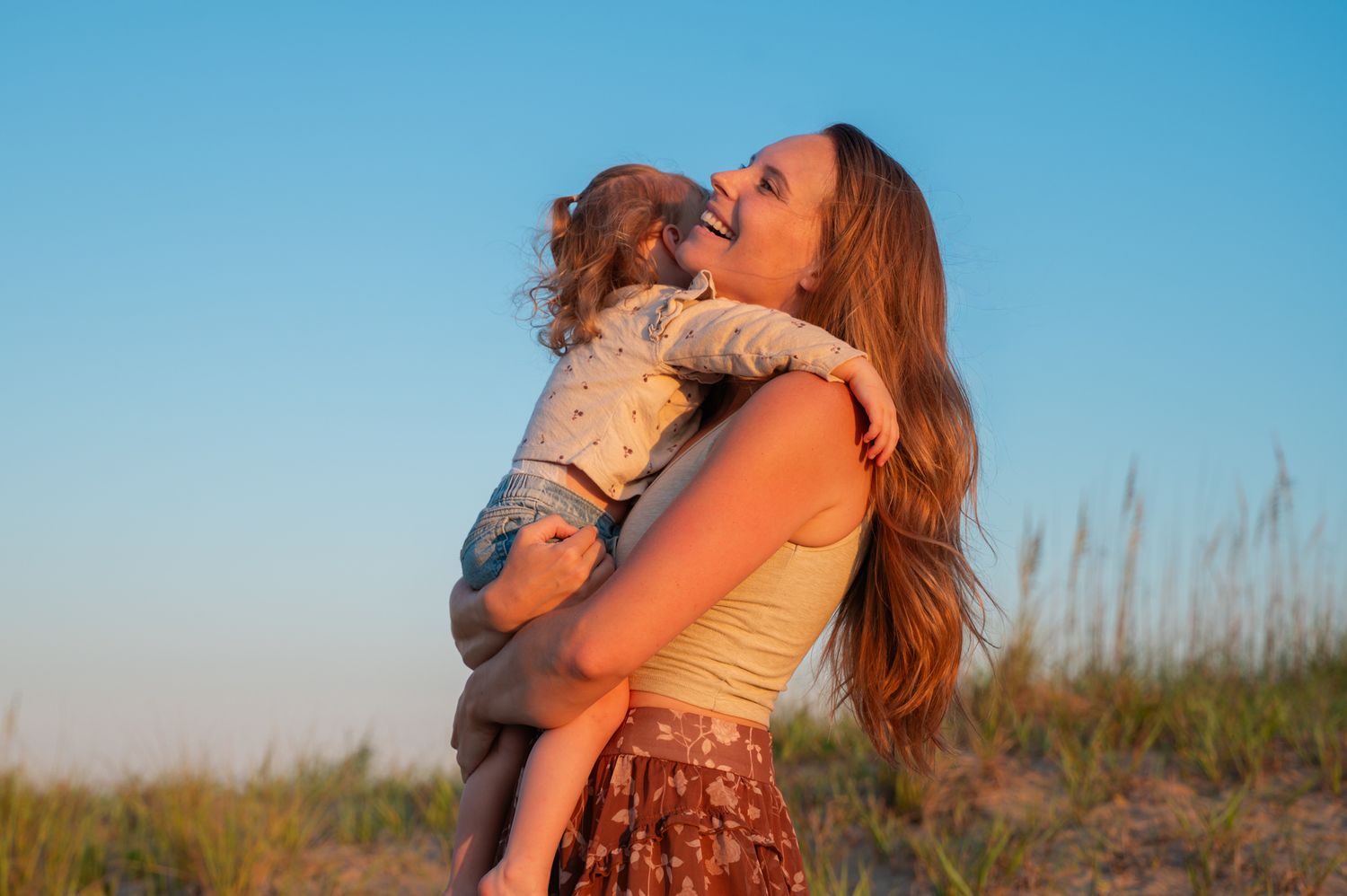 Laughing mother holding her young child against a clear blue sky at a sandy beach.