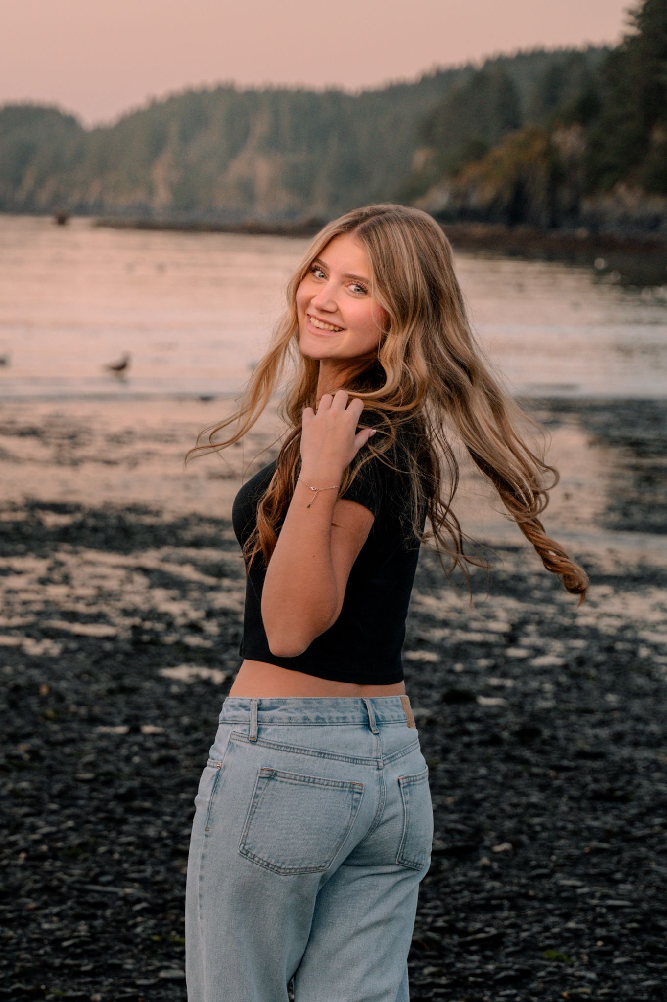 Young woman with long blonde hair in black crop top and jeans smiling on a rocky beach at sunset.
