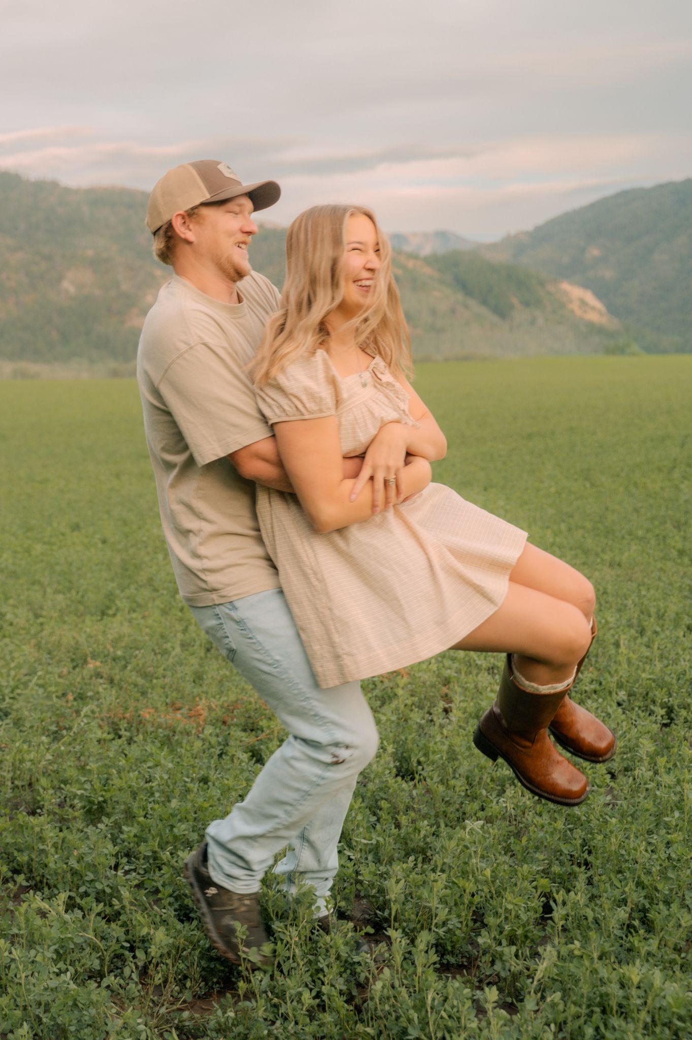 Happy couple laughing in a green field at golden hour, man lifting woman in beige dress and brown boots.