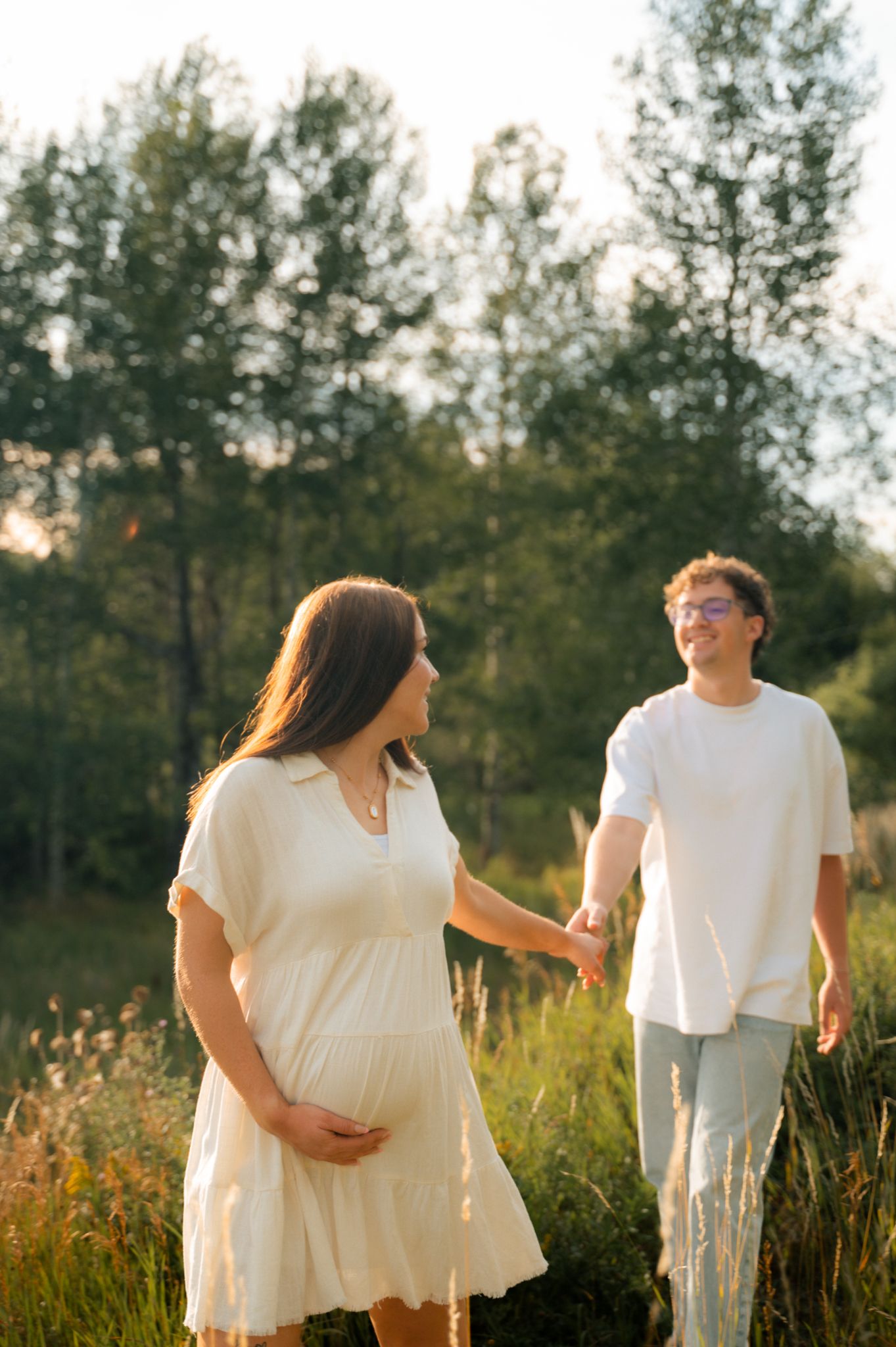 Pregnant couple holding hands walking through sunlit meadow at golden hour.
