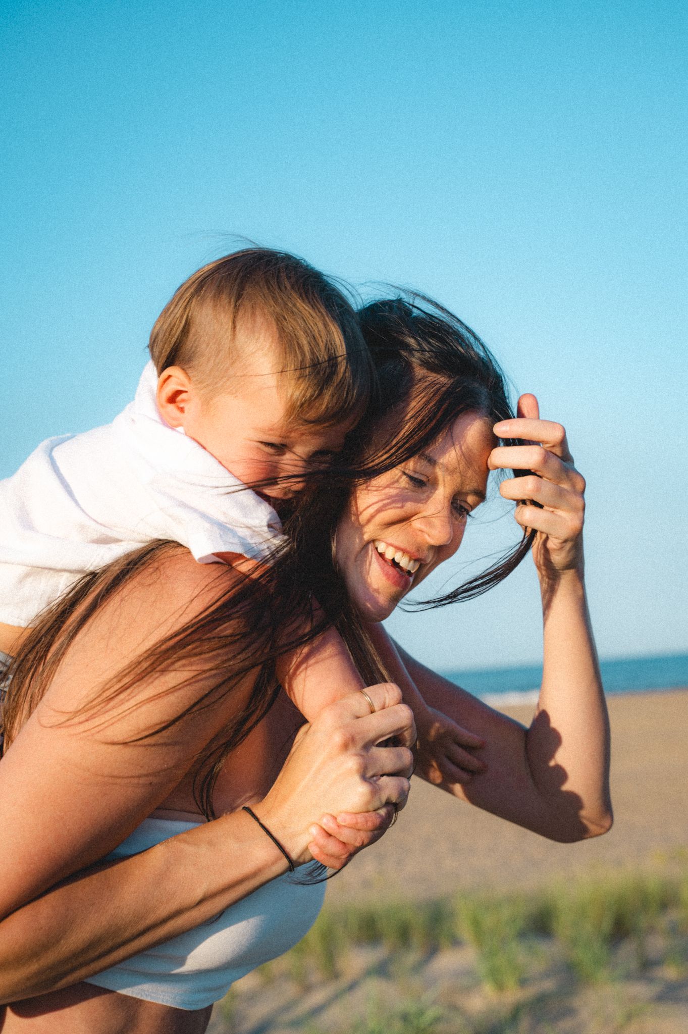 Joyful mother carrying laughing baby on her back at a sunny beach.