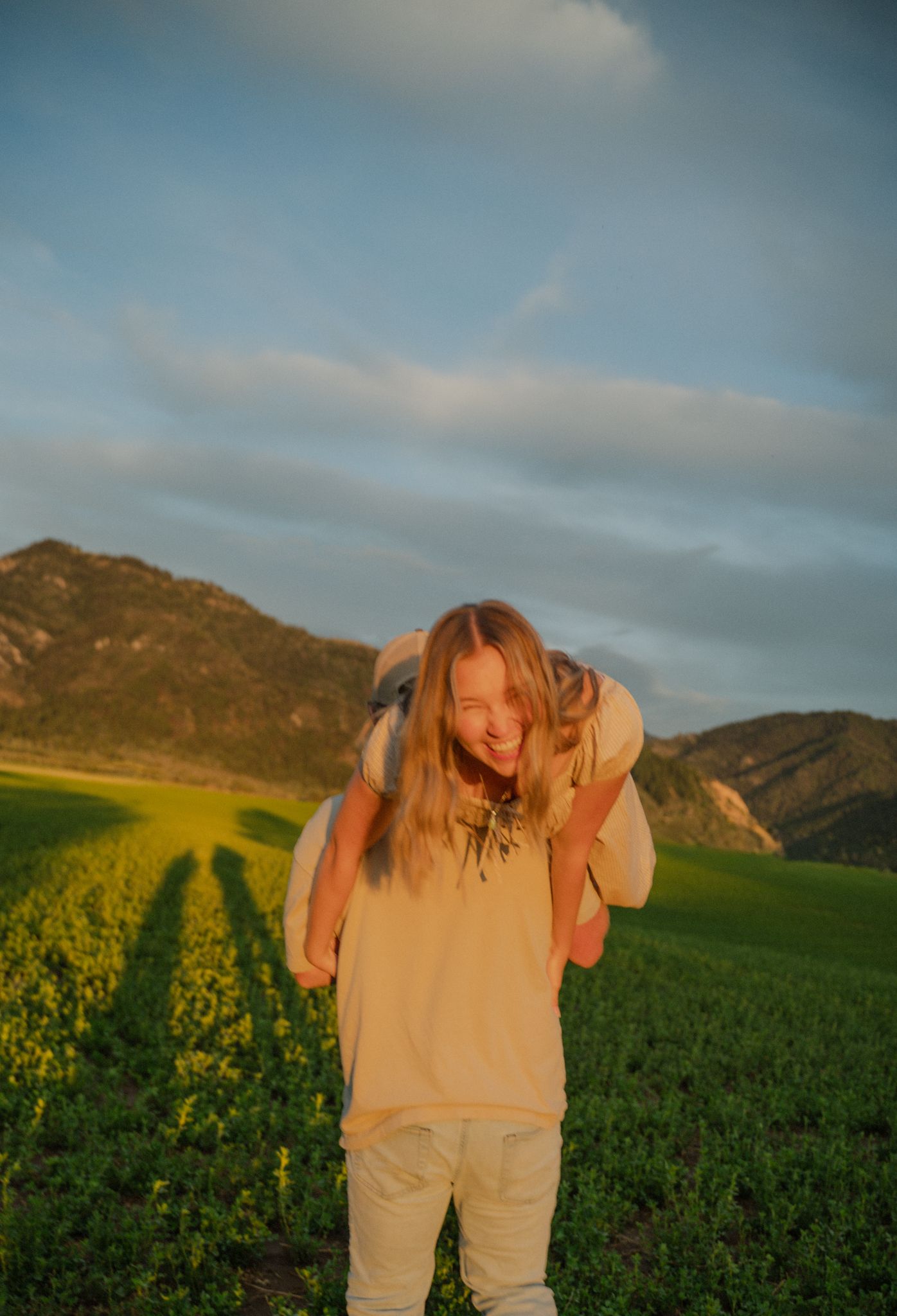 Person carrying someone on their back laughing in a green field with mountain backdrop.