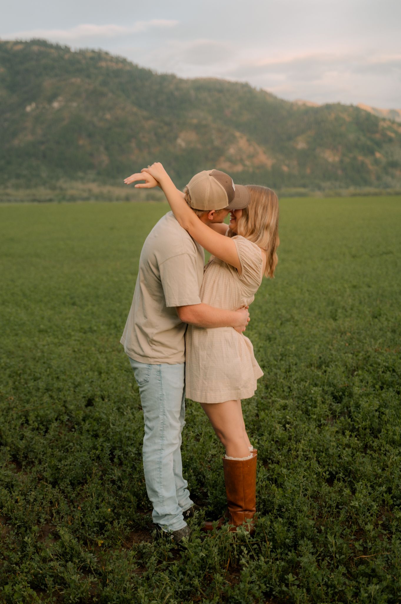 Romantic couple kissing and embracing in a green field with a mountain backdrop during golden hour.