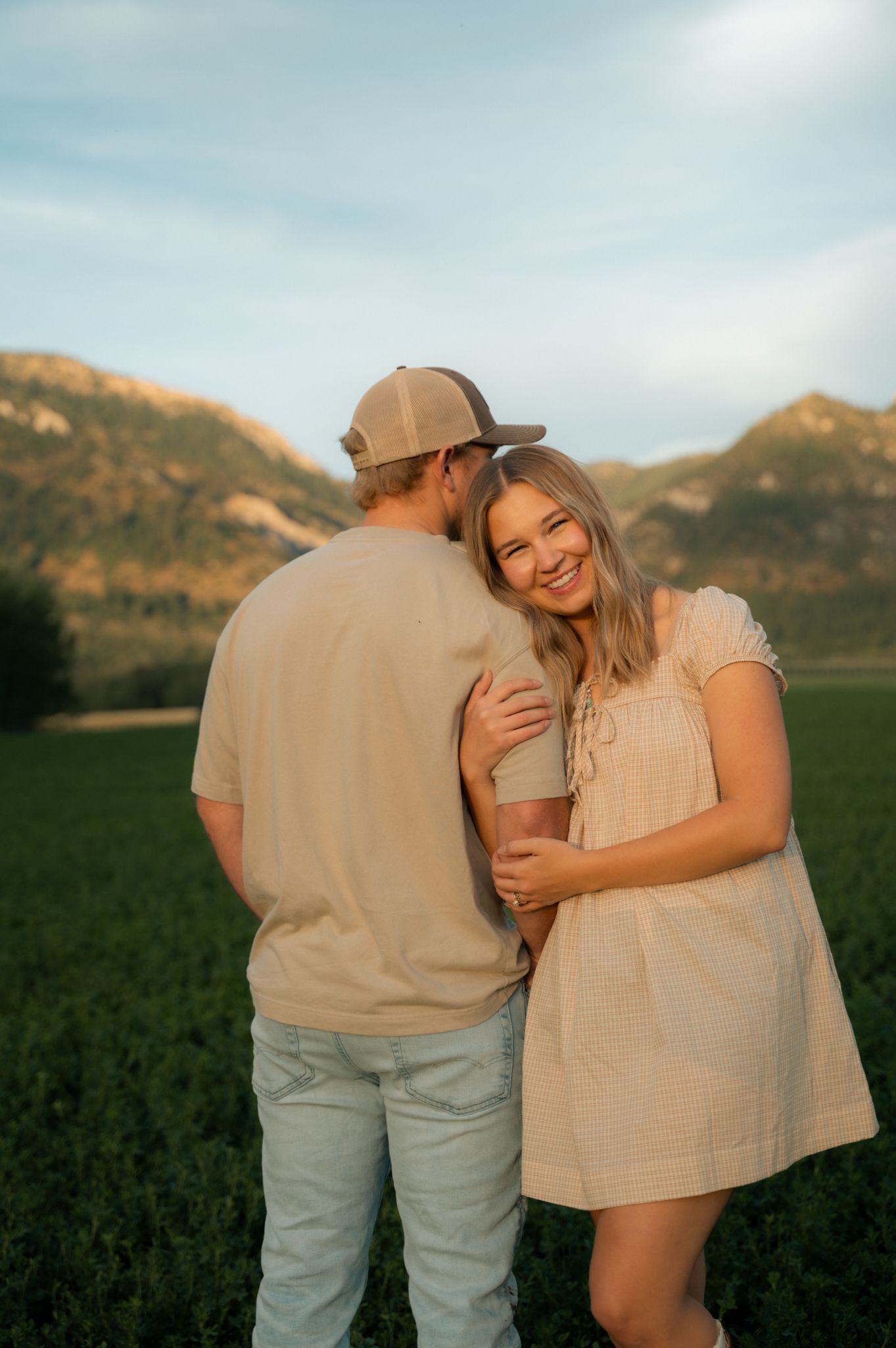 Smiling young woman in cream dress embraces man in tan cap in a sunlit green field.