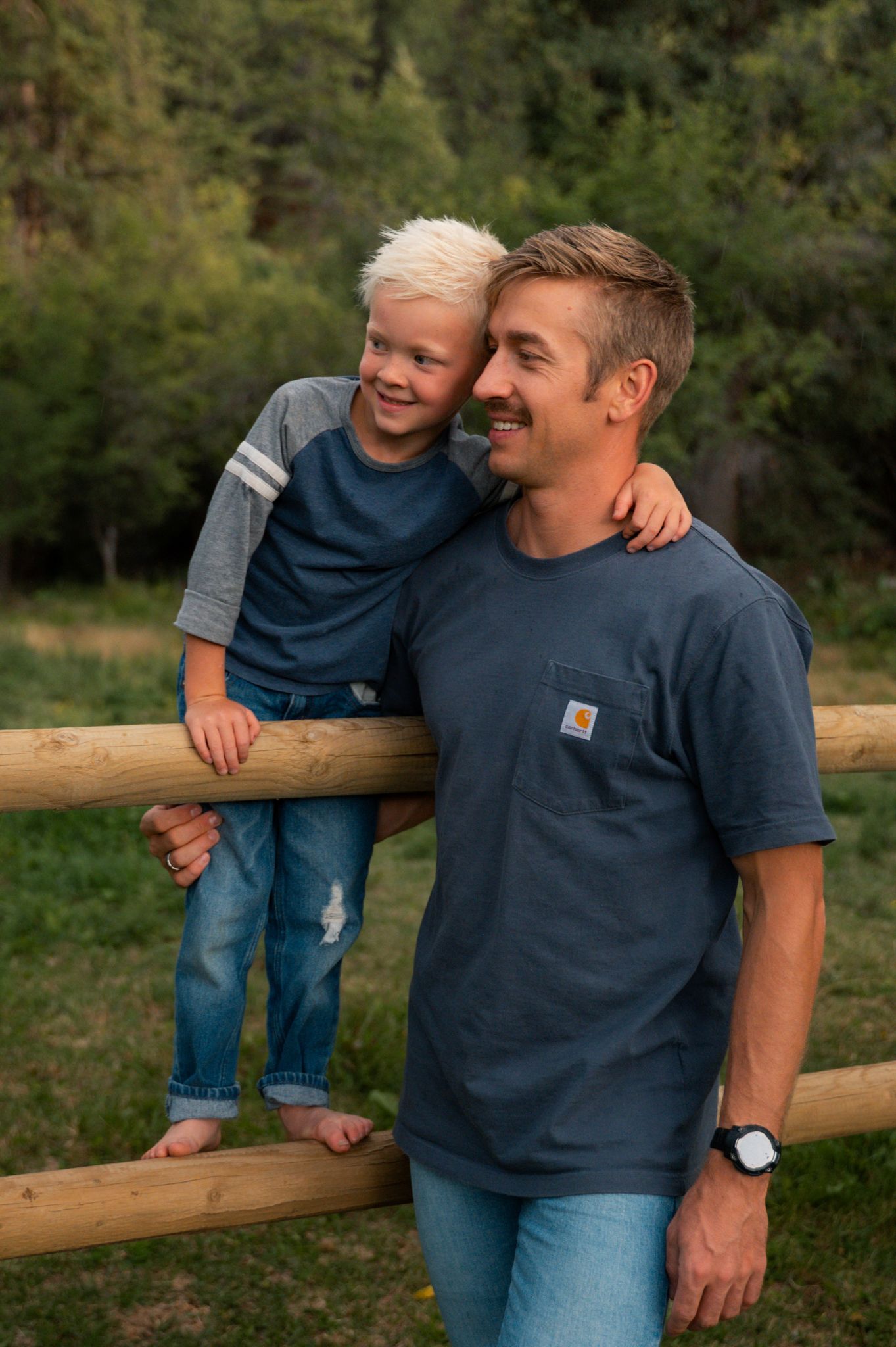 Smiling boy sitting on wooden fence leaning on man in navy Carhartt tee in a green outdoor setting.