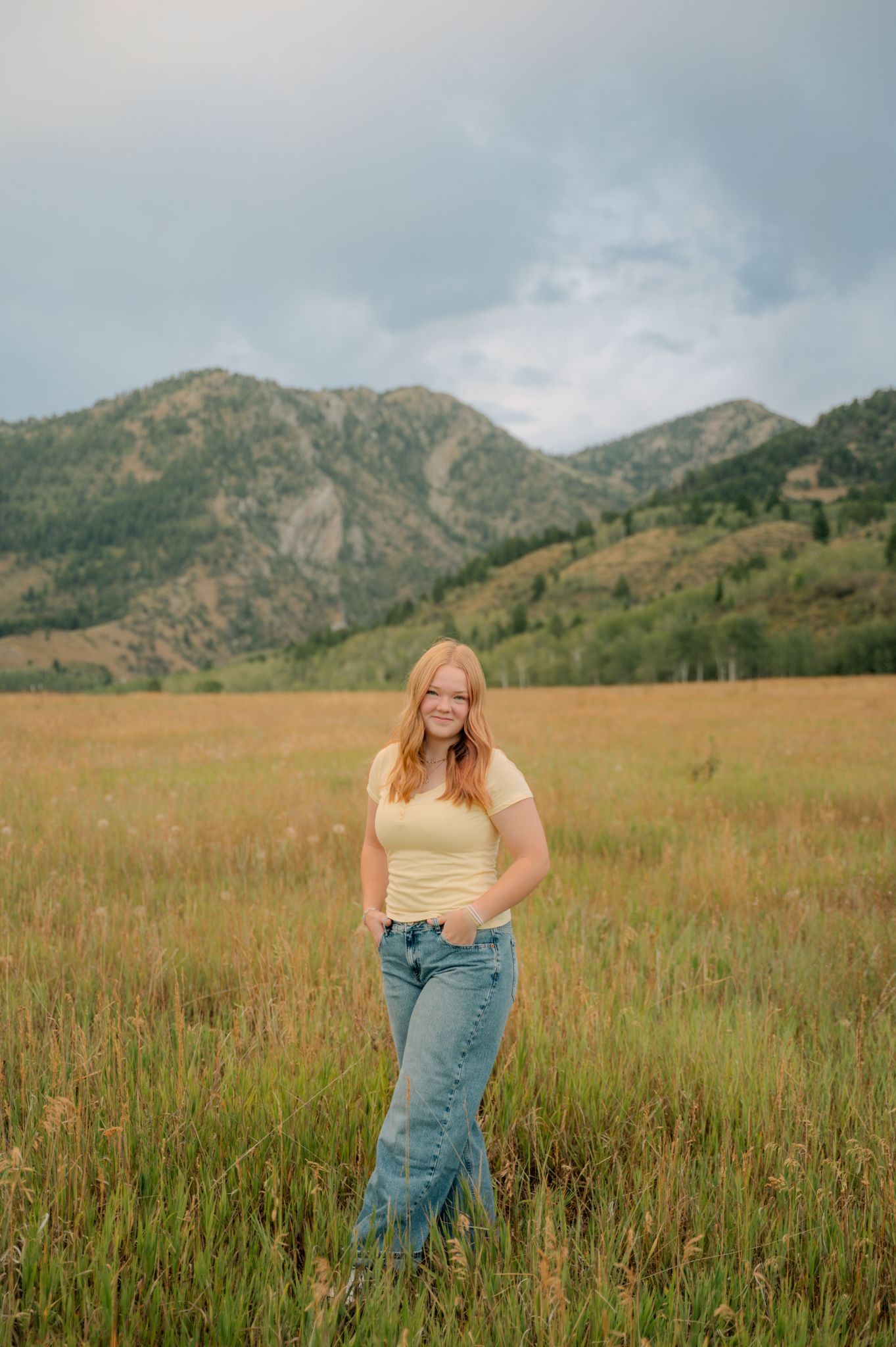A red-haired teenage girl in a yellow top and jeans stands in a field with mountain backdrop.