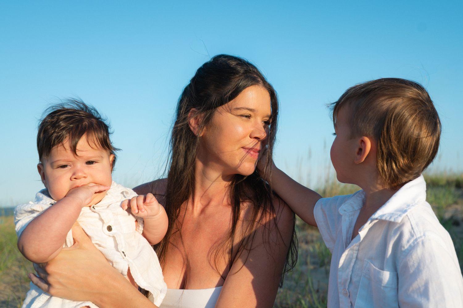 Mother with dark hair holding infant while toddler stands beside her outdoors.