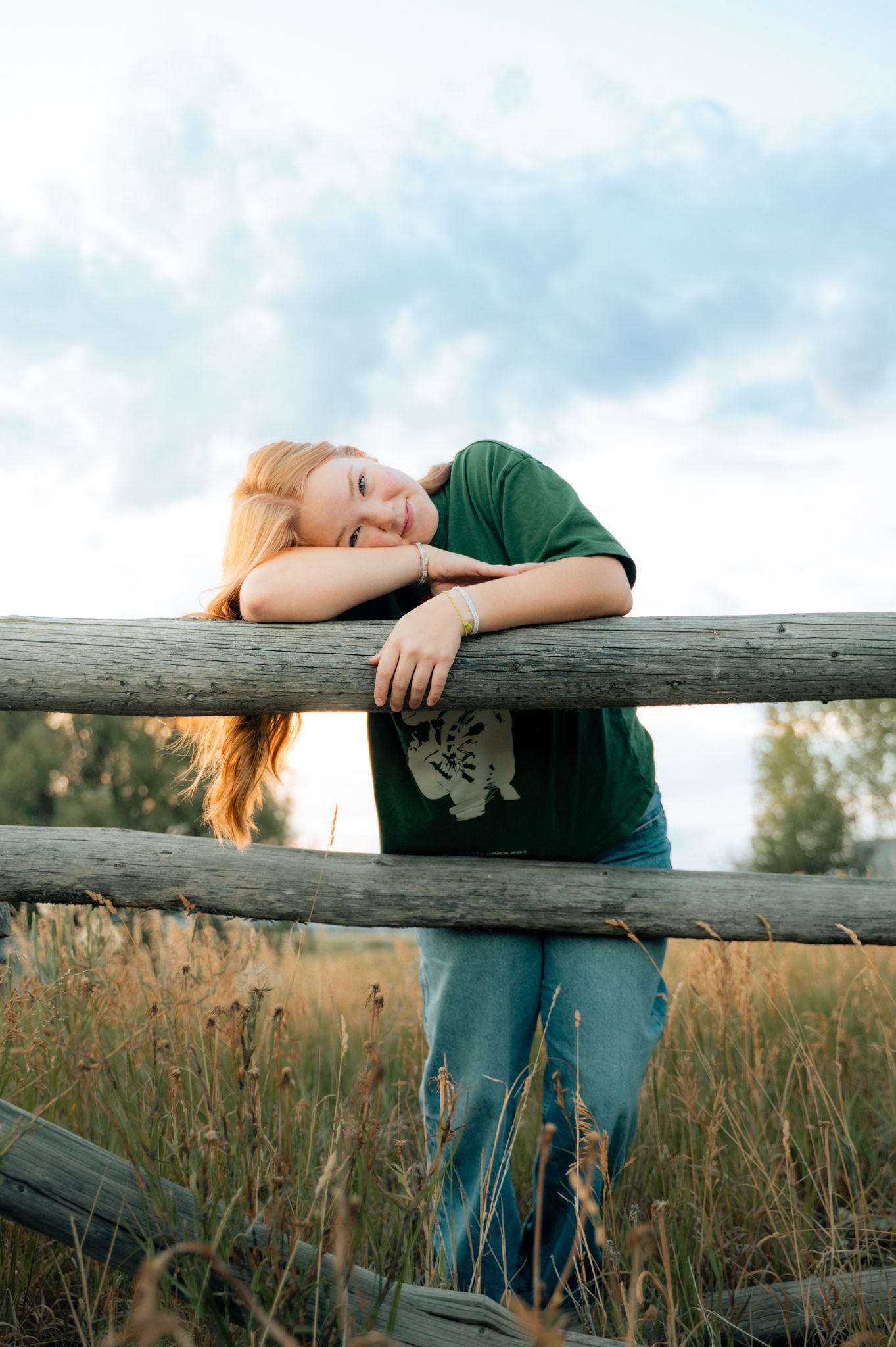 Young woman with long red hair resting on a rustic wooden fence in a field.