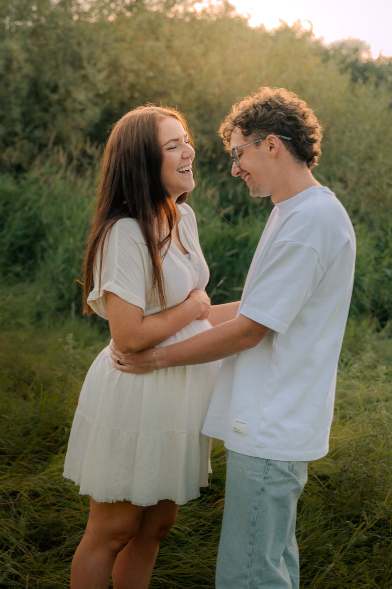 Happy young couple laughing and embracing in a lush green field at golden hour sunset.