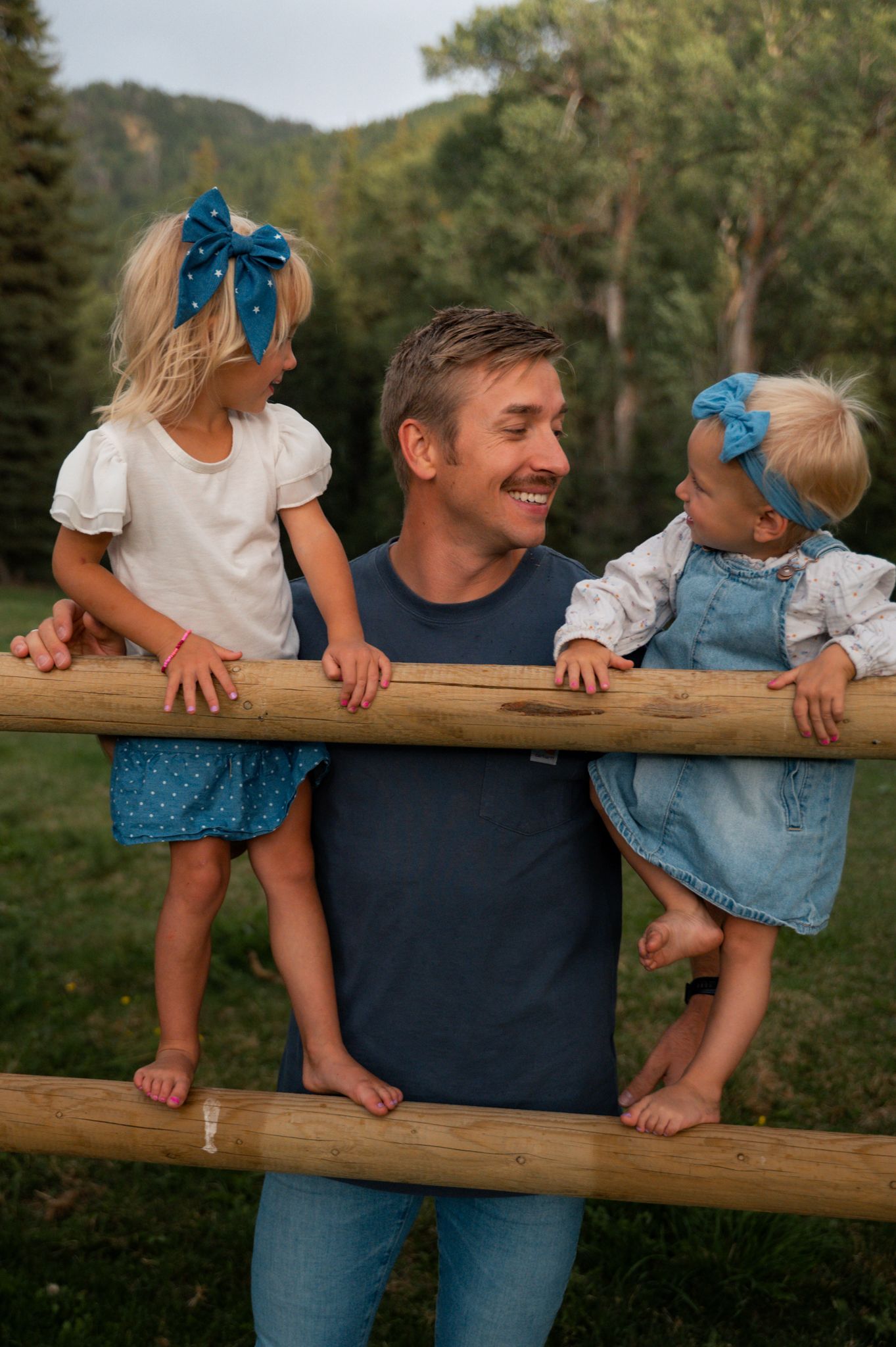 Happy father leans on wooden fence while two young blonde daughters in blue bows smile at him.