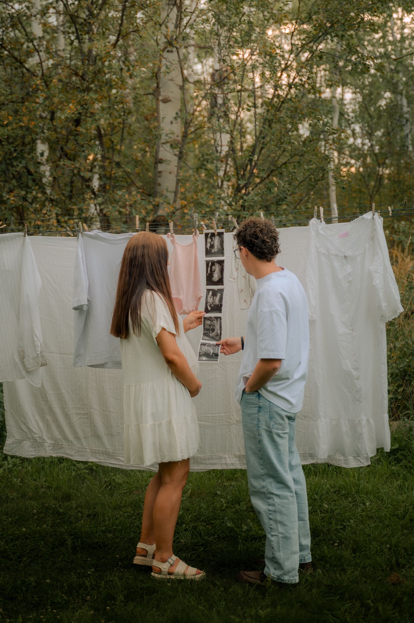Couple looking at ultrasound photos hanging on a clothesline with white laundry in a wooded backyard.
