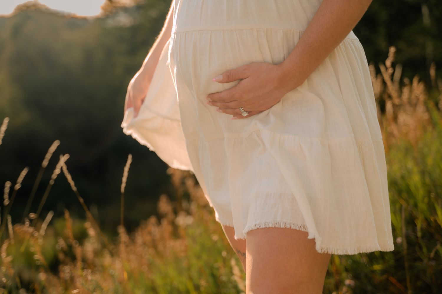 Pregnant woman in white dress cradling baby bump in golden sunlit meadow field.