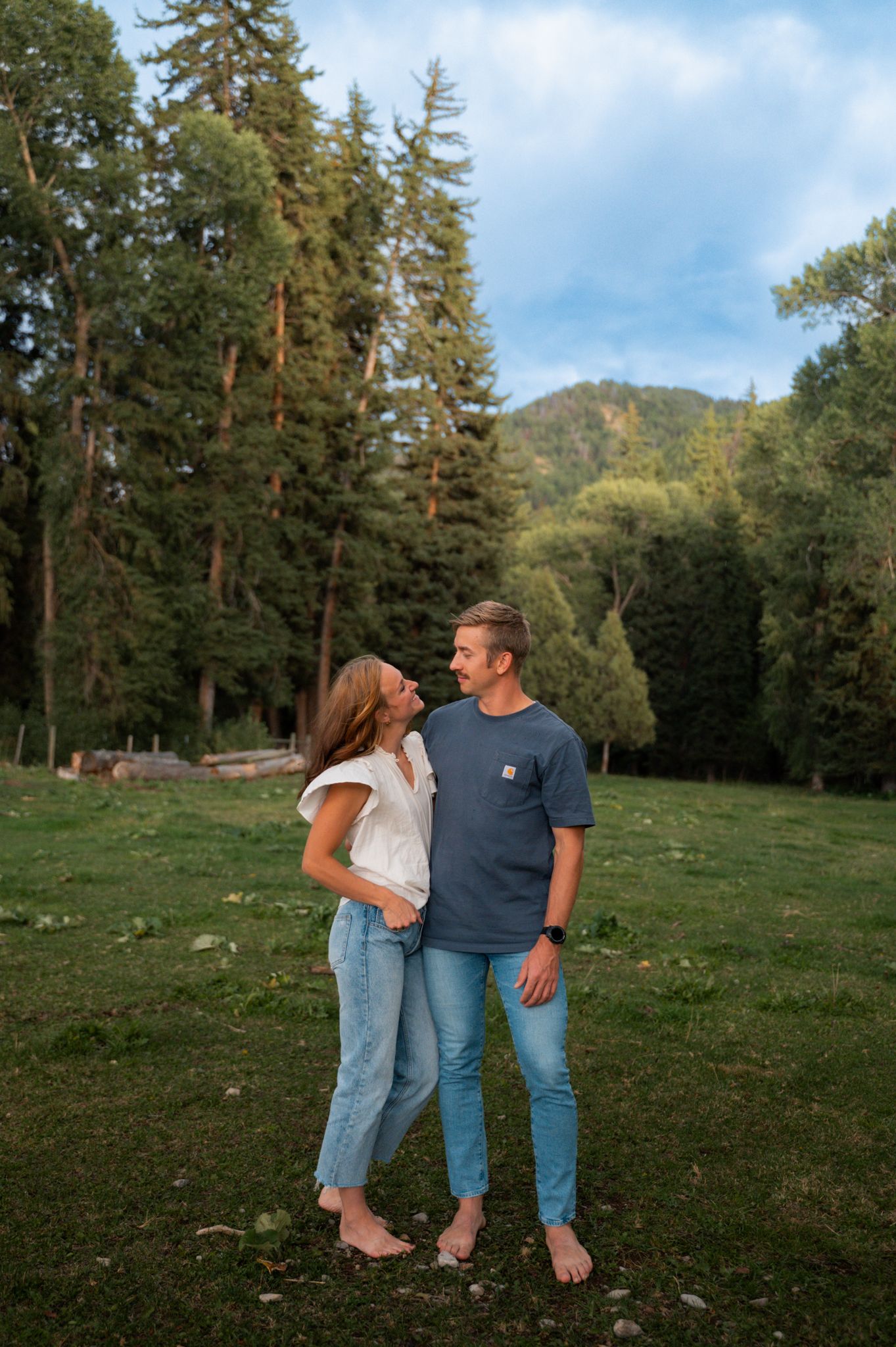 Couple smiling at each other barefoot on green grass with tall pine trees and mountain backdrop.