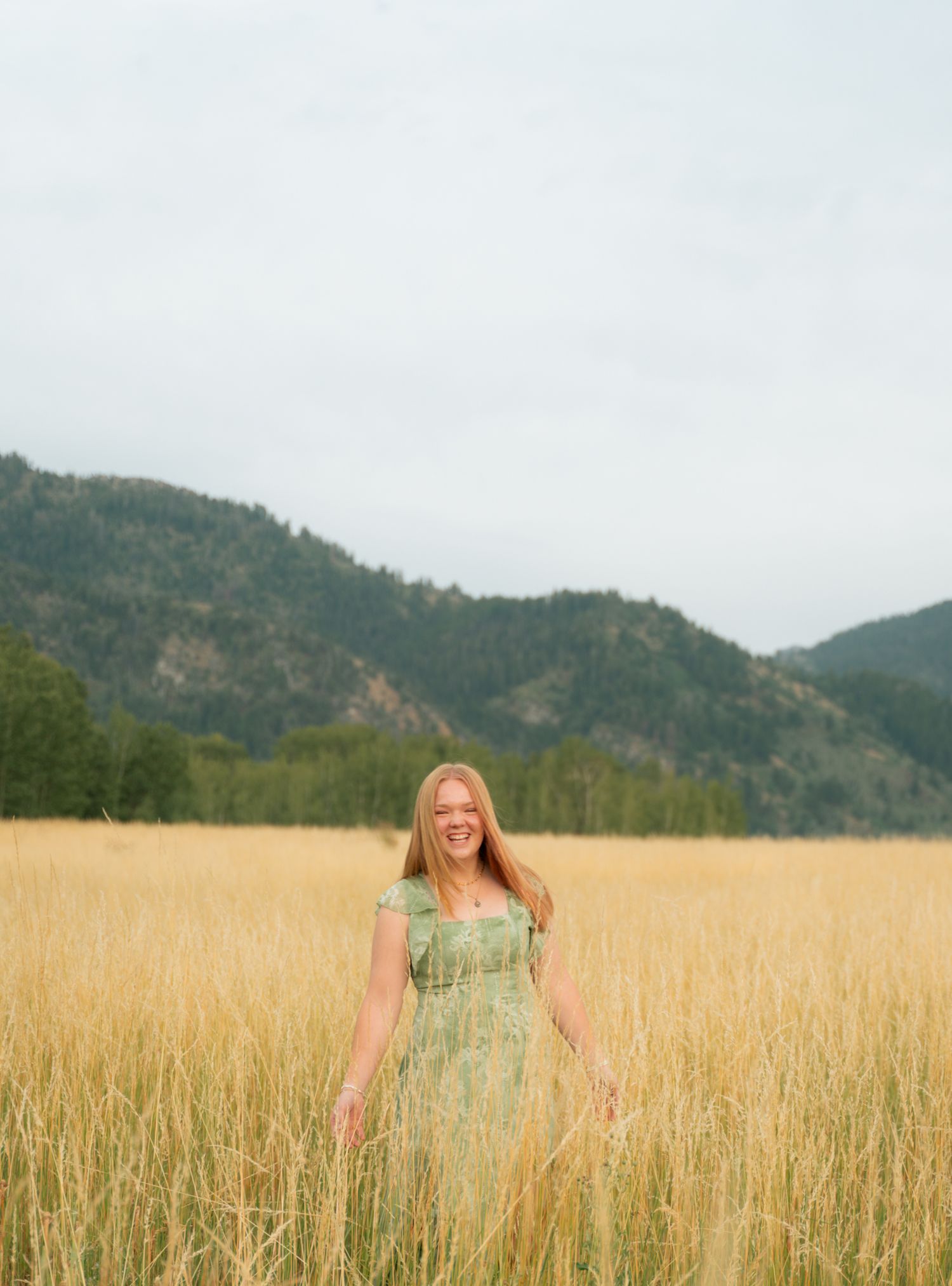 A smiling redhead woman in a green floral dress stands in a golden grass field with mountains.