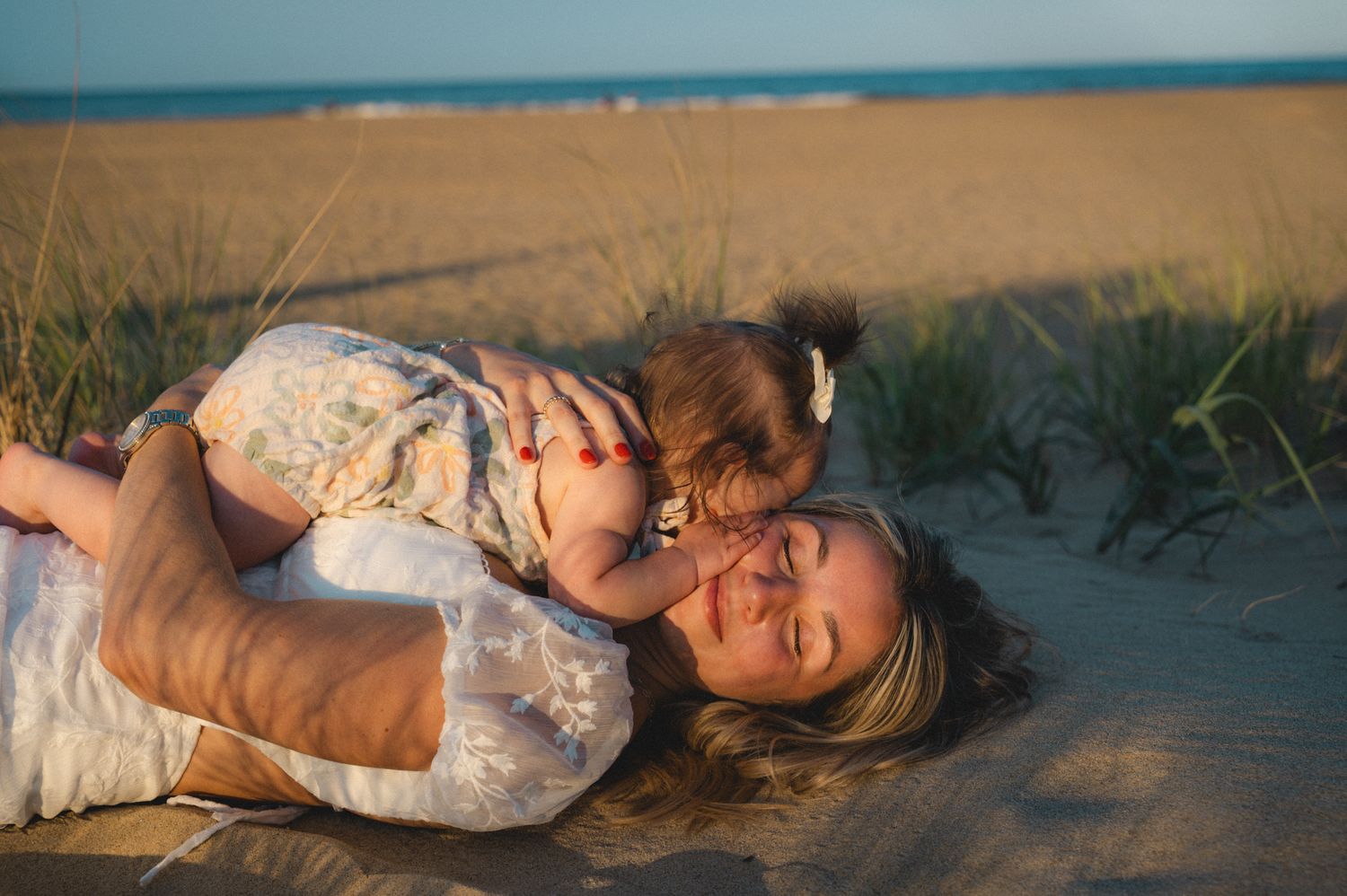 Mother lying on sandy beach dunes cuddling her baby in golden sunset light.