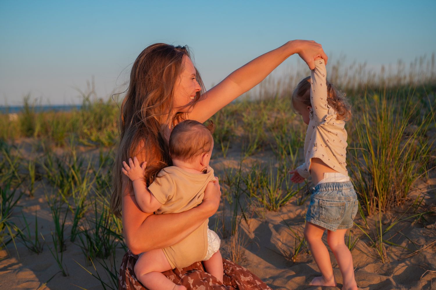 Mother sitting on beach sand holding infant while toddler stands nearby at sunset.