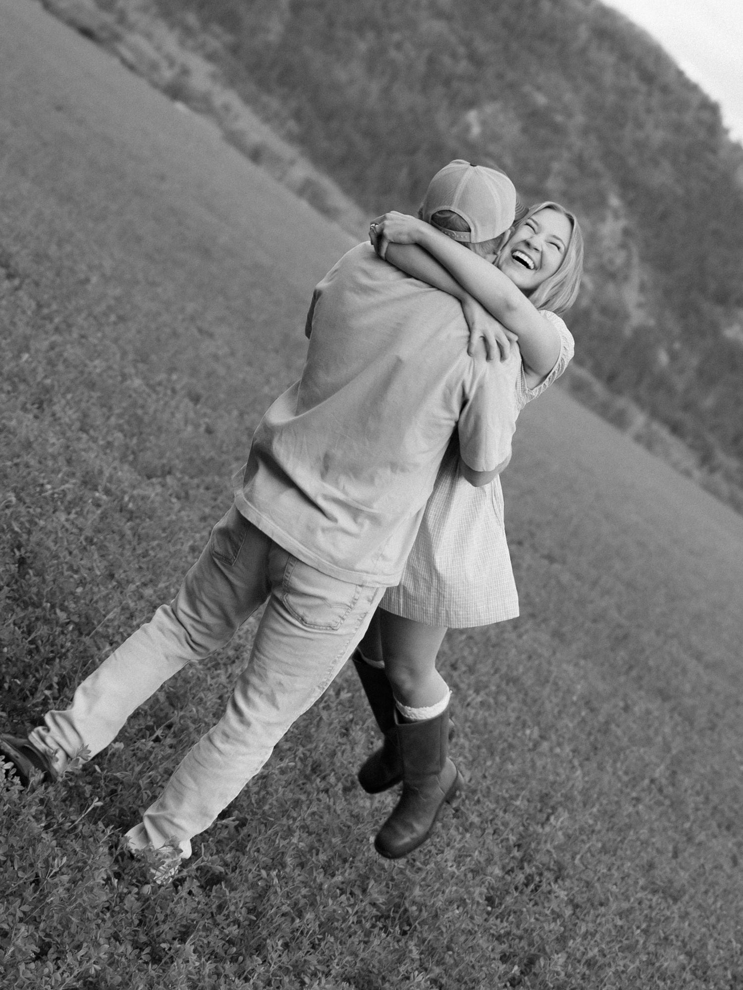 A joyful couple embraces and laughs in a black and white photo in an open green field.