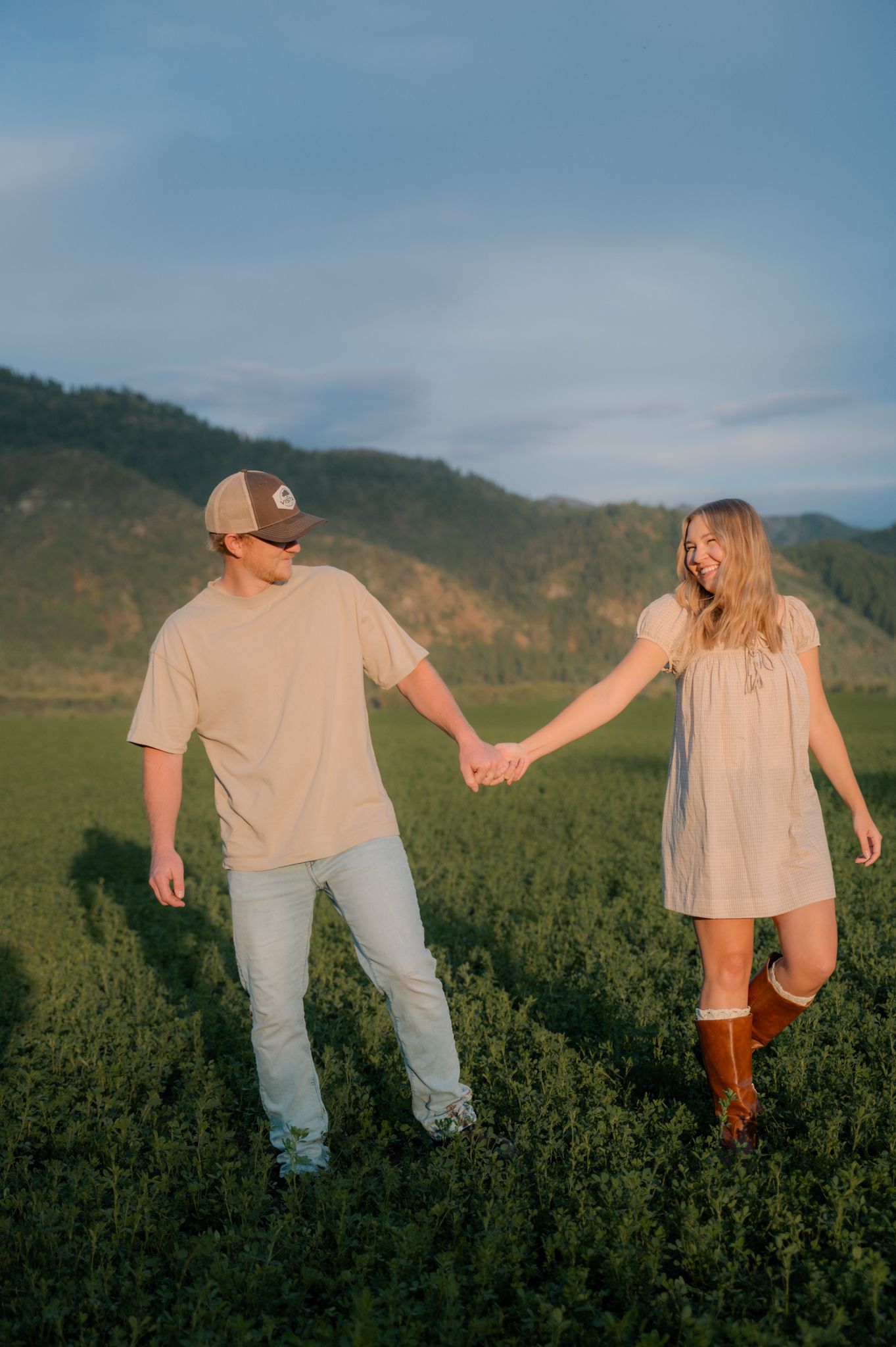 Couple holding hands and walking through a lush green field with mountain scenery in the background.