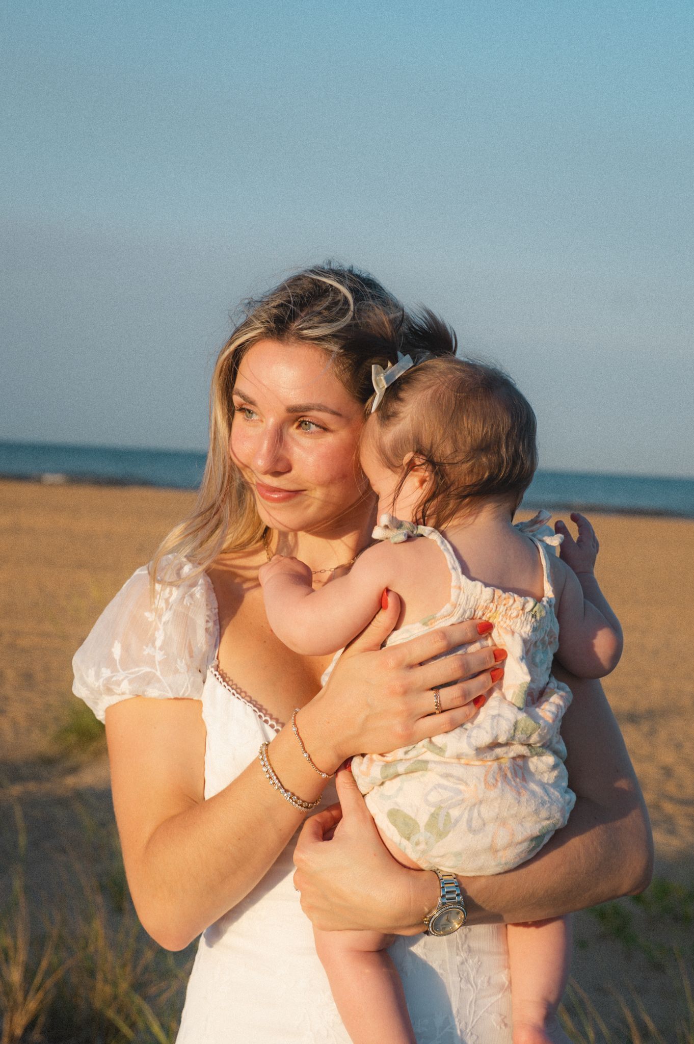 Woman in white dress holding baby on sunny beach at golden hour.