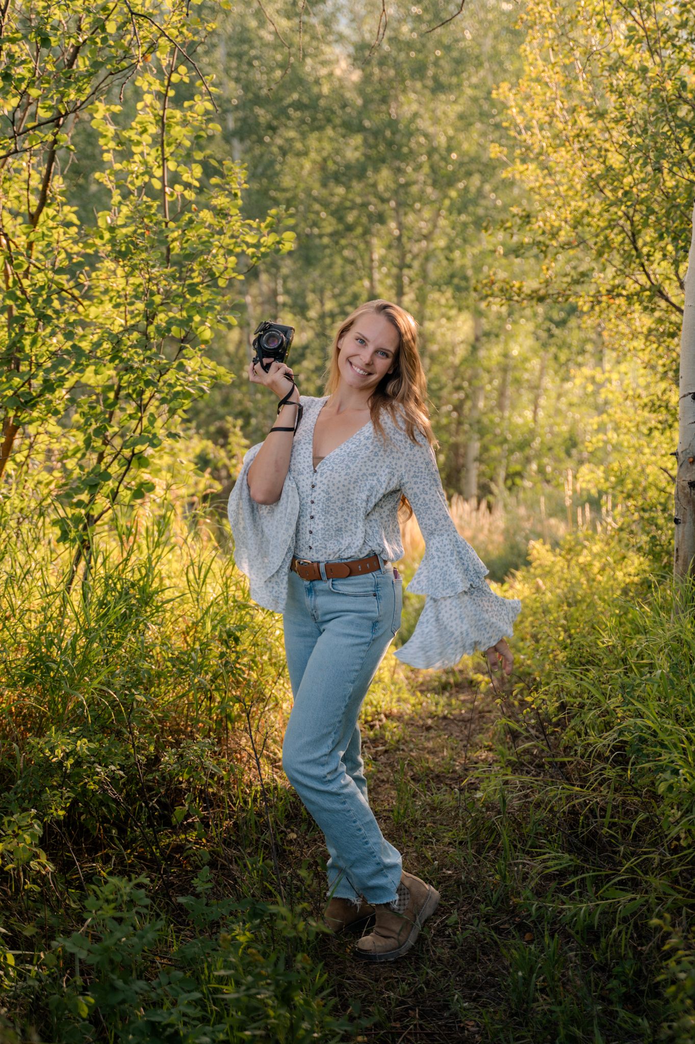 A smiling blonde female photographer poses with her camera in a sunlit forest of golden aspen trees.