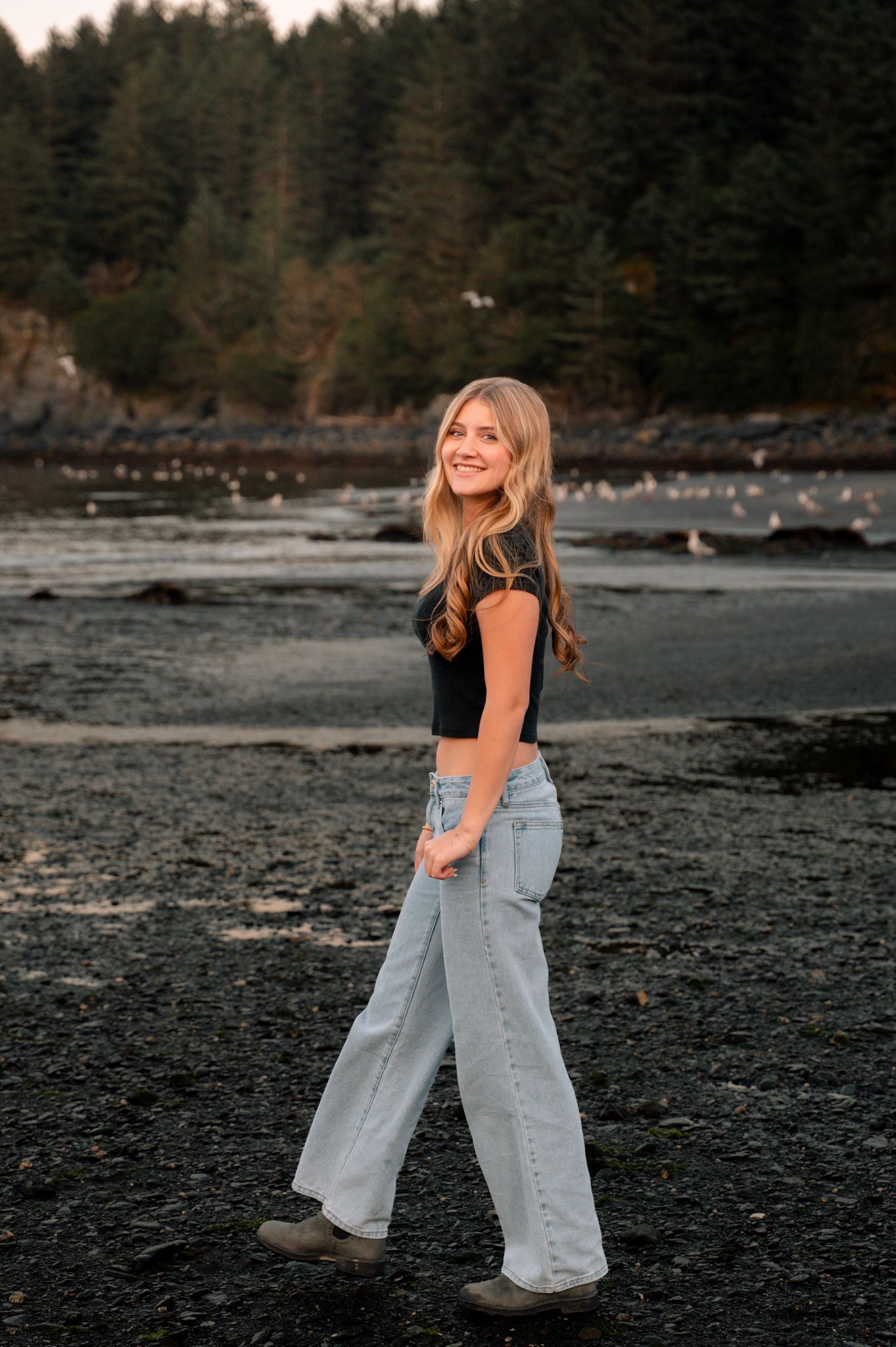 Young woman in black top and wide-leg jeans smiling on a rocky beach with evergreen trees behind her.