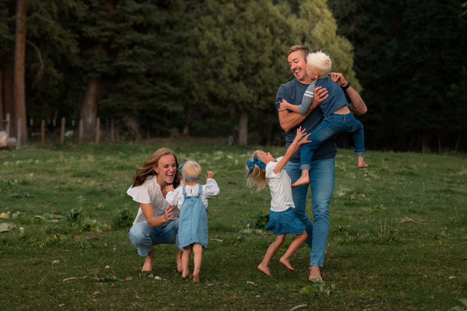 Joyful family playing together on green grass in a pine forest, parents lifting and hugging young children.