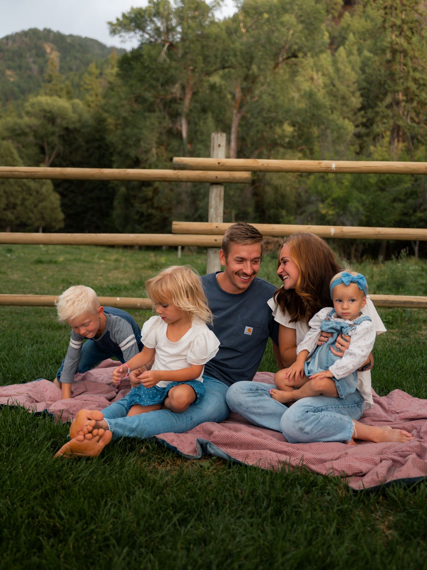 Happy family of five sitting on pink blanket in green meadow with mountain backdrop.