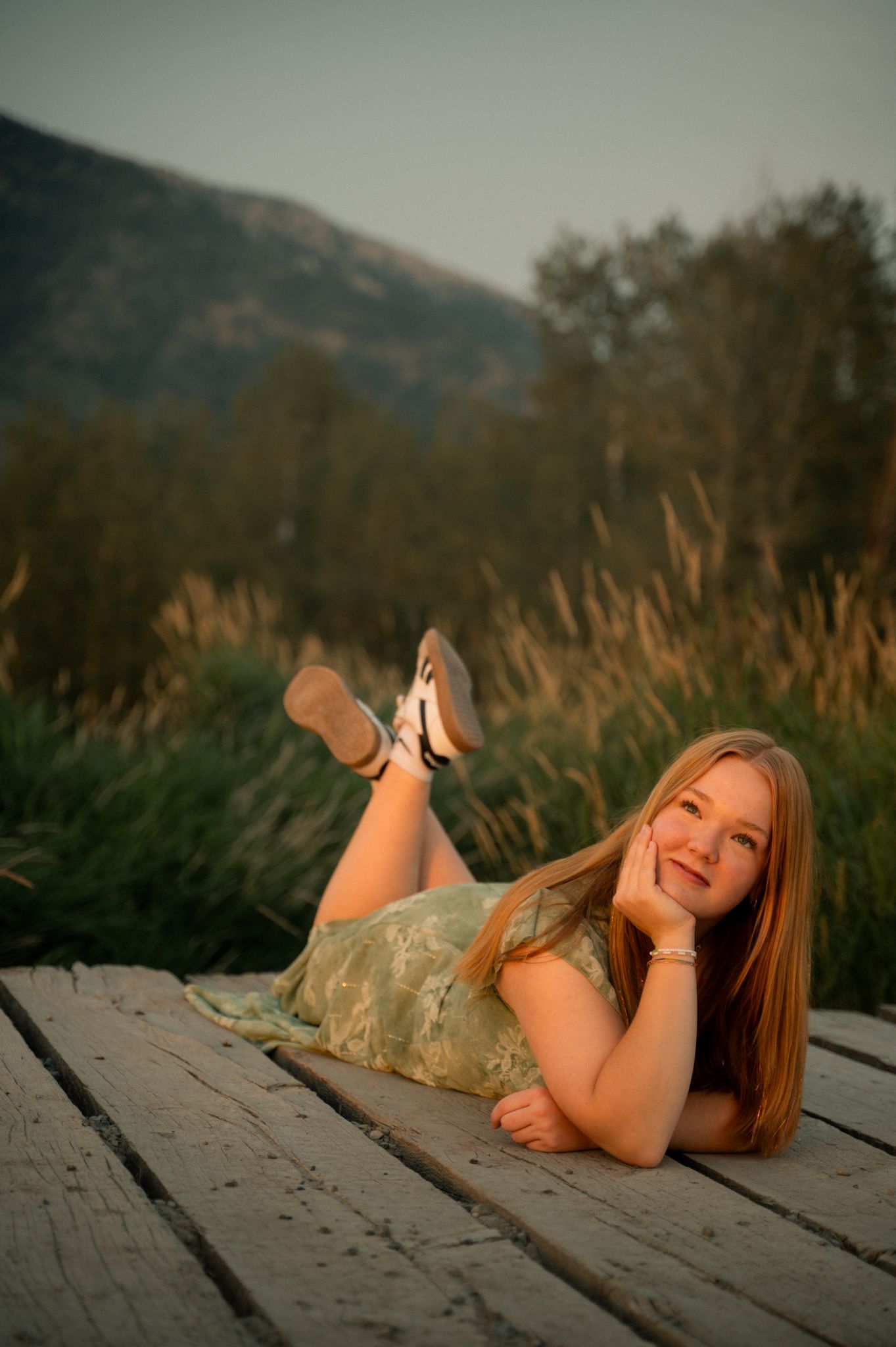 Teen girl in green dress lying on a wooden dock smiling, with mountains in background.