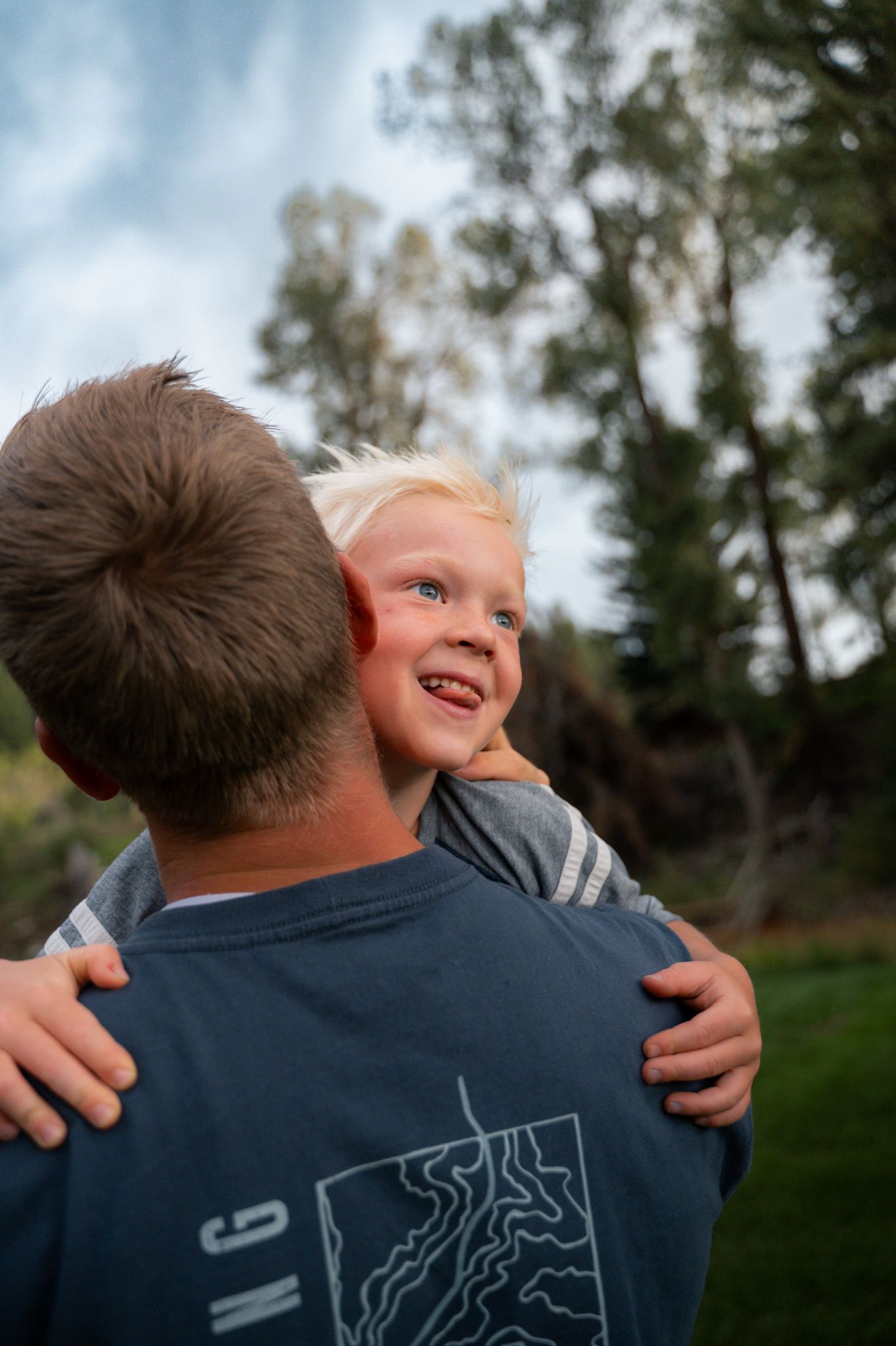 Smiling blonde toddler being held by father outdoors among tall pine trees.
