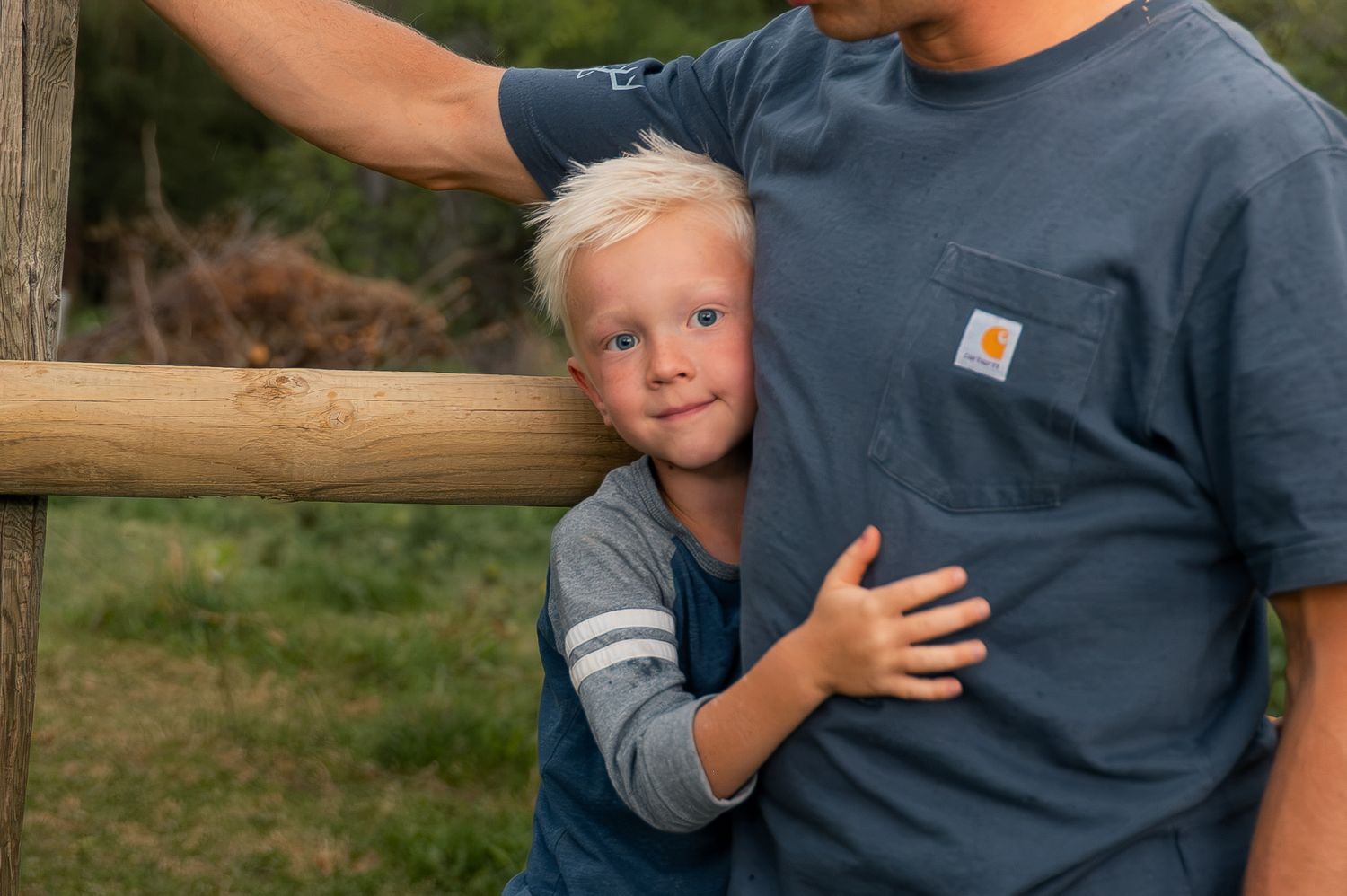 Young blonde boy hugging his father near a rustic wooden fence outdoors.